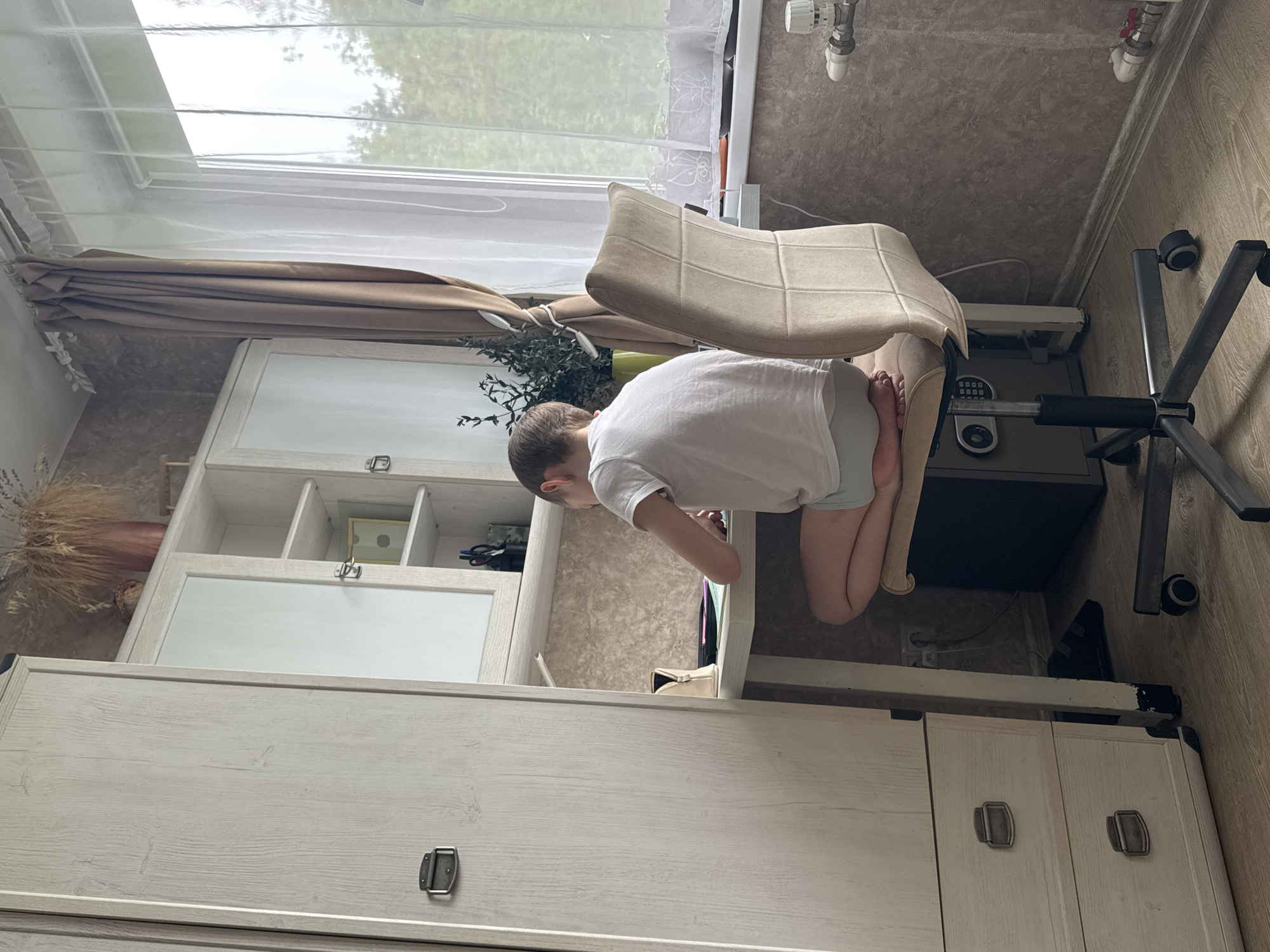 A child in a white shirt is bent over a partially open drawer of a white cabinet. The child is positioned near an office chair. A window with a beige curtain is visible in the upper left corner of the image. The setting appears to be indoors. Раздолье, Russia is not explicitly visible, but inferred as the location.
