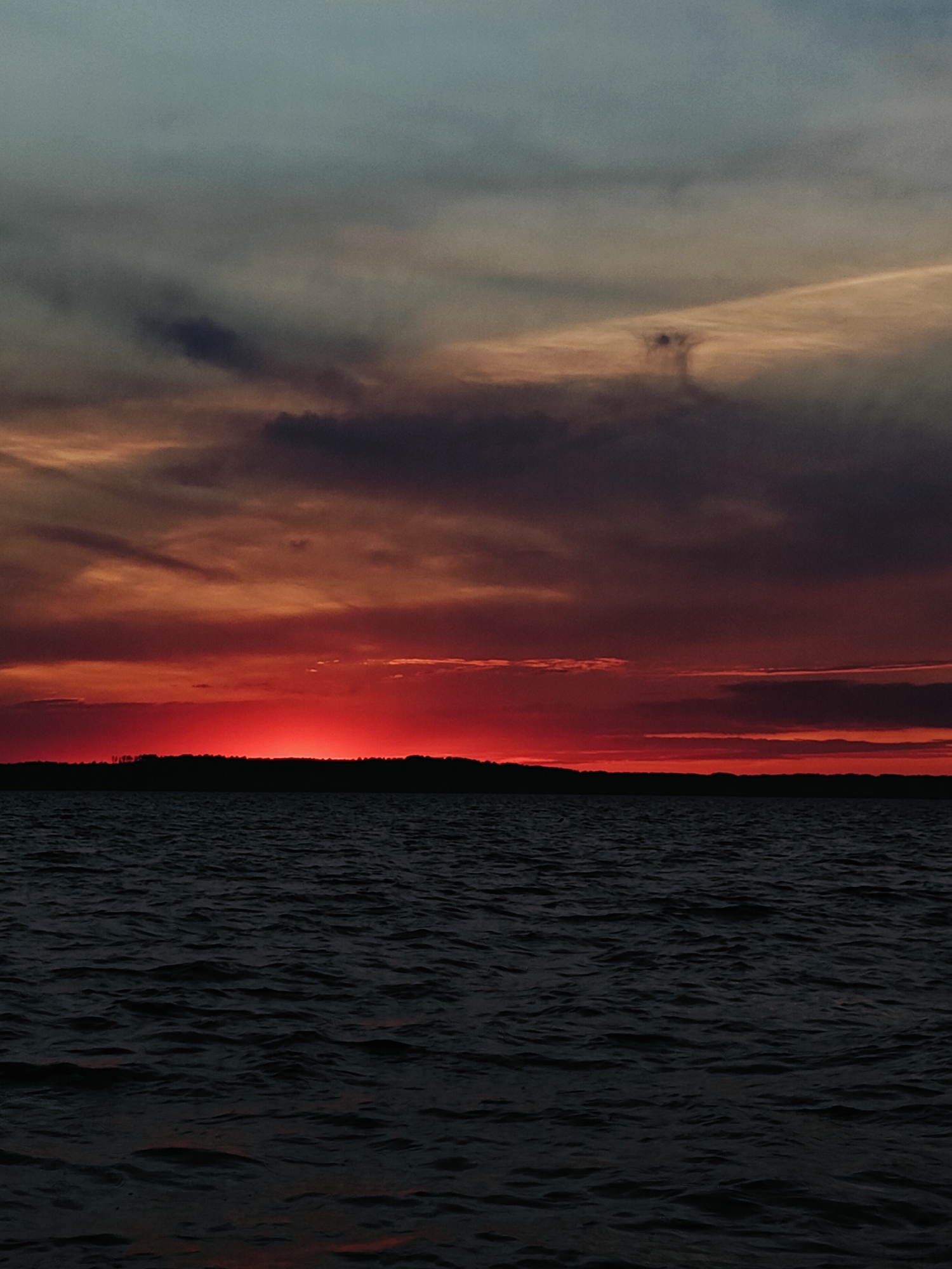 The image shows a sunset over a body of water. The sky is filled with dramatic clouds in shades of red, orange, purple, and gray. A dark, silhouetted landmass, possibly a forest or hills, forms a distinct horizon line between the sky and the water. The water itself is dark and choppy, reflecting the colors of the sunset in muted tones. The overall impression is one of a moody and intense natural scene. The specific location in Stepanovo, Russia is not discernible from the image itself.