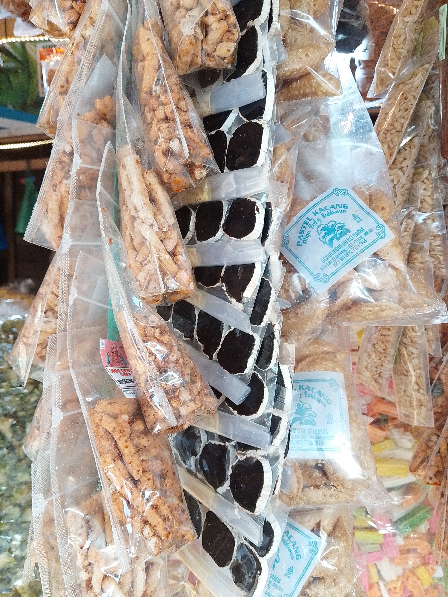 This image shows a close-up view of numerous plastic bags filled with various snacks, likely displayed for sale. The snacks appear to be a mixture of fried goods and possibly candies or nuts. Some bags contain light-brown, irregularly shaped fried items, while others hold what look like whole peanuts or other small, elongated snacks. There are also some black and white items visible in a different packaging, appearing as circular pieces with white edges.
Several labels are visible on some of the bags, written in Indonesian. One prominent label reads "PASTEL KACANG RABBANA" and beneath it "USAHA INDUSTRI KELUARGA". Another label, partially visible, says "UMMI DEWI SNACK BASRENG".
The overall presentation suggests a market stall or small shop, typical of local commerce in Indonesia. The lighting is somewhat dim, with some bright highlights on the plastic packaging, suggesting artificial lighting or sunlight filtering through. The background is blurred but hints at more merchandise or a shop interior. The image is taken at a street-level perspective. The location is identified as Sawang, Indonesia, implying this is a typical scene of food retail in that region.