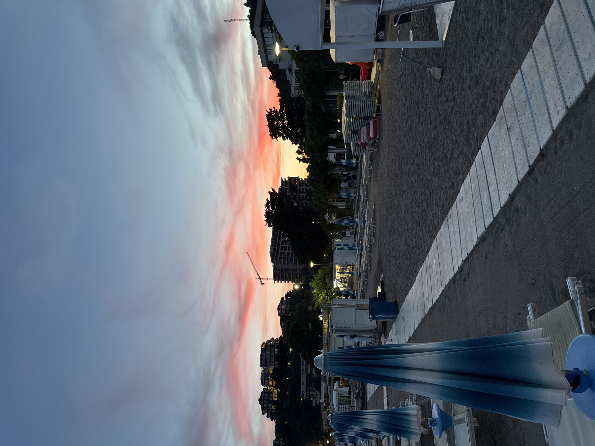 This image captures a sunset scene on a beach in Boreti, Montenegro. The sky is painted with vibrant hues of pink and orange, transitioning to a clear blue at the top. In the foreground, several closed beach umbrellas with blue and white stripes stand upright on the dark sand. A white wooden walkway extends into the distance, leading past rows of neatly stacked lounge chairs. In the mid-ground, small buildings, possibly changing rooms or snack bars, are visible. Further in the background, a crane and several apartment buildings are situated on a hill overlooking the beach. A few people are scattered across the beach, some walking and others appearing to be socializing. The overall atmosphere is serene and tranquil, indicative of the end of a day at the seaside.