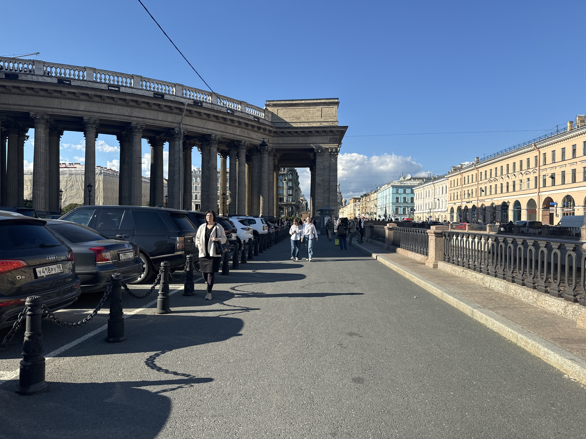 The image captures a street scene in Saint Petersburg, Russia, likely on a sunny day. On the left, a grand, colonnaded building, characteristic of neoclassical architecture, dominates the foreground. This structure is identified as the Kazan Cathedral. Cars are parked along the sidewalk in front of the cathedral, separated by black bollards and chains. Several people are walking on the sidewalk and crossing the street. On the right side of the image, a row of colorful buildings with arched storefronts lines the street, indicating a commercial area. A decorative iron fence runs along the edge of the sidewalk, separating the street from what appears to be a canal or river. The sky is clear blue with a few scattered white clouds. The overall impression is one of an active urban environment with historical architecture.