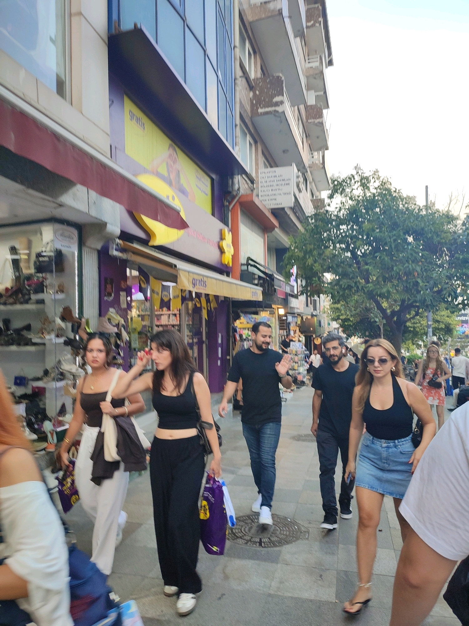 The image depicts a street scene in Kadıköy, Turkey, with a focus on pedestrians walking along a sidewalk lined with shops. In the foreground, a group of people are walking towards the camera. To the left, a woman in a dark tank top and white wide-leg pants is holding a light-colored tote bag. Next to her, a woman in a black tank top and black wide-leg pants is carrying purple shopping bags. Further ahead, two men are walking. The man on the left is wearing a black t-shirt and jeans and is waving. The man to his right is wearing a black t-shirt and dark pants. To the right of these men, a woman in a black tank top and a denim skirt is walking, wearing sunglasses. Behind her, a woman in a floral dress is visible.
The shops lining the street have awnings and display windows. One shop on the left has shelves filled with shoes. Above the shops, buildings with balconies are visible. A tree with green foliage is situated on the right side of the street. The overall environment suggests a commercial area with active pedestrian traffic. The lighting indicates it is daytime.