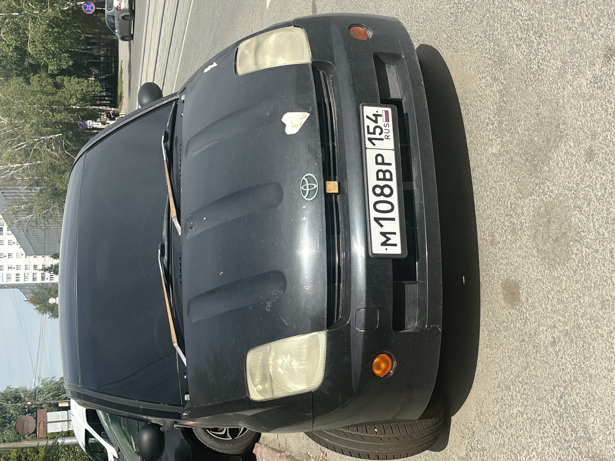 A black Toyota Prius is parked on an asphalt road in Novosibirsk, Russia. The car has tinted windows and a white heart-shaped sticker on the hood. The license plate reads "M108BP 154 RUS". In the background, there are trees, buildings, and a tram track. The sky is clear and the sun is shining.