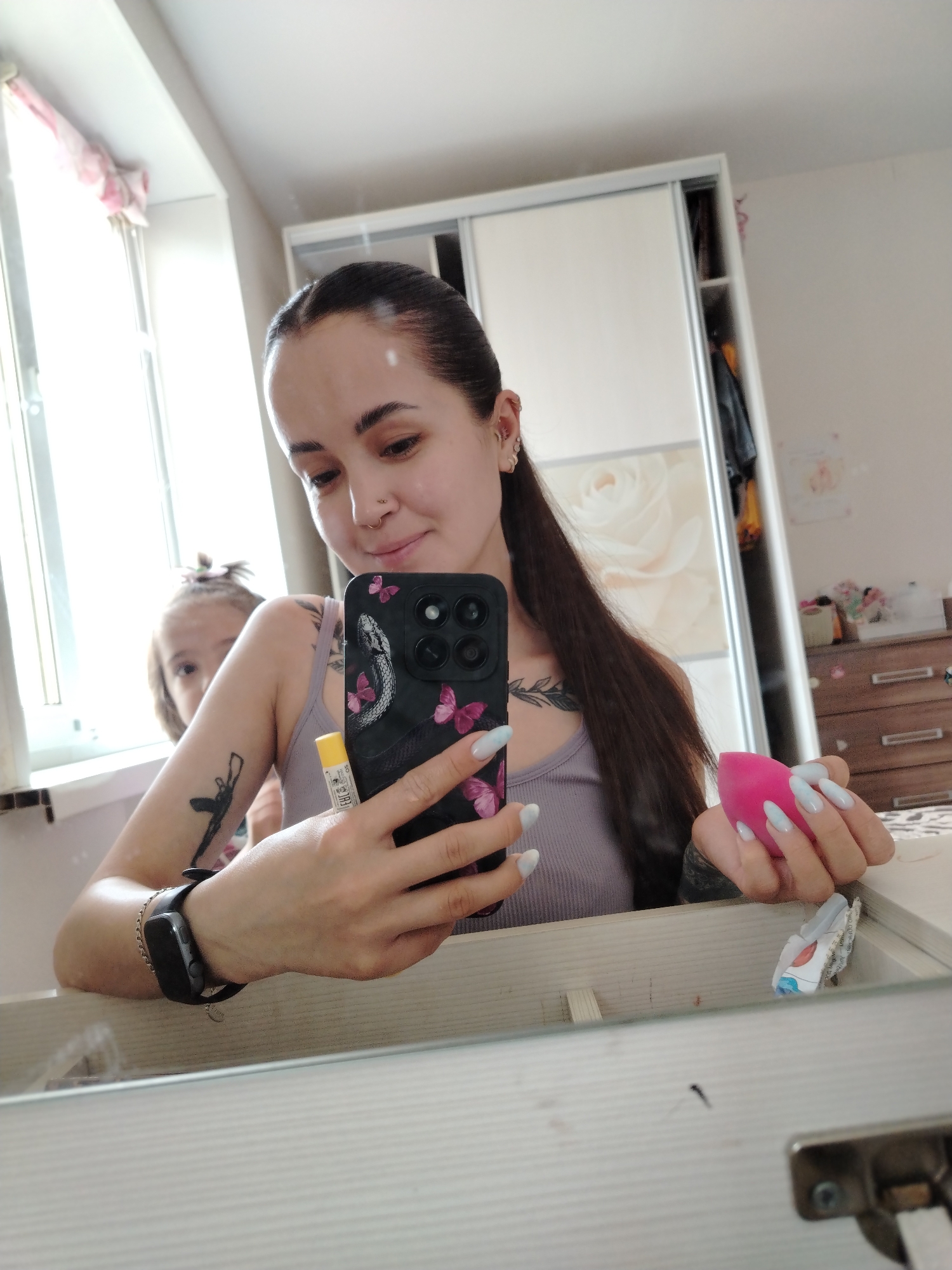 A woman is taking a selfie in a room. She is holding a phone in her right hand and a pink makeup sponge in her left. She has a tattoo on her left arm and collarbone. She is wearing a black smartwatch. In the background, there is a child near the window and a white wardrobe. There is also a dresser with decorative objects on it. The location is likely a domestic interior. Noviy, Russia is not evident.