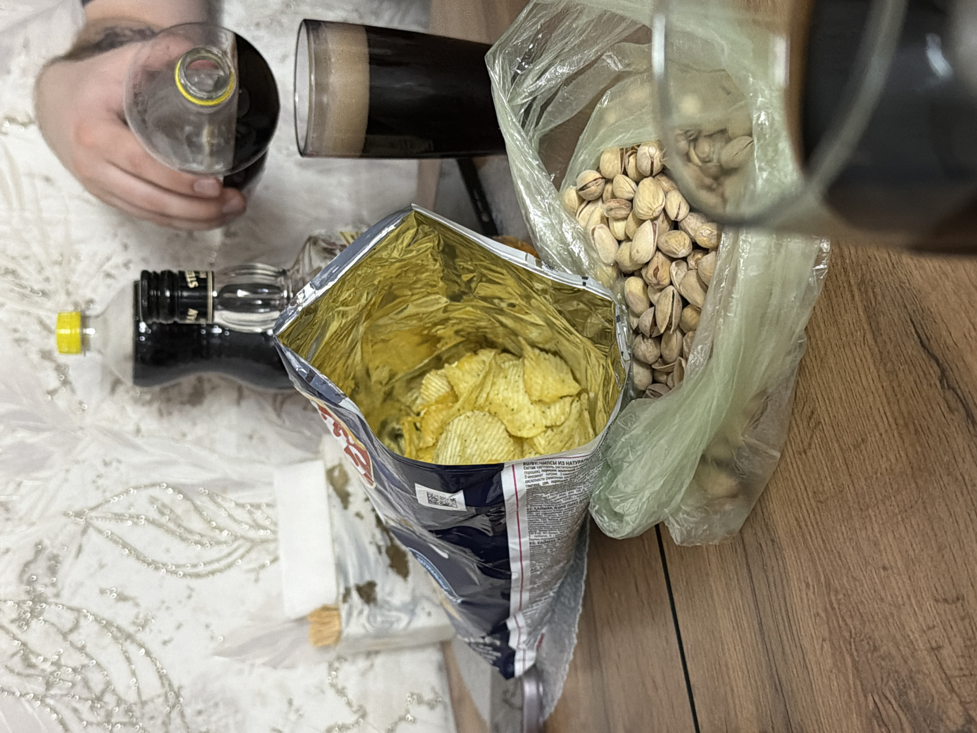 A cluttered still life is presented, featuring a variety of consumables. Two glass bottles and a pint glass of dark liquid, likely beer, are arranged amongst the food. The bag of potato chips has a shiny gold inner lining and is filled with ruffled yellow chips. Pistachios are contained in a clear plastic bag. The scene is set on a surface of two different types. A wooden-patterned surface on the right contrasts with a white fabric or similar material with an embroidered pattern on the left. The upper left corner shows a human hand holding one of the glass bottles. The scene is indicative of an informal gathering or a personal indulgence. This image appears to have been taken in Russia, in the city of Reshetnikovo.