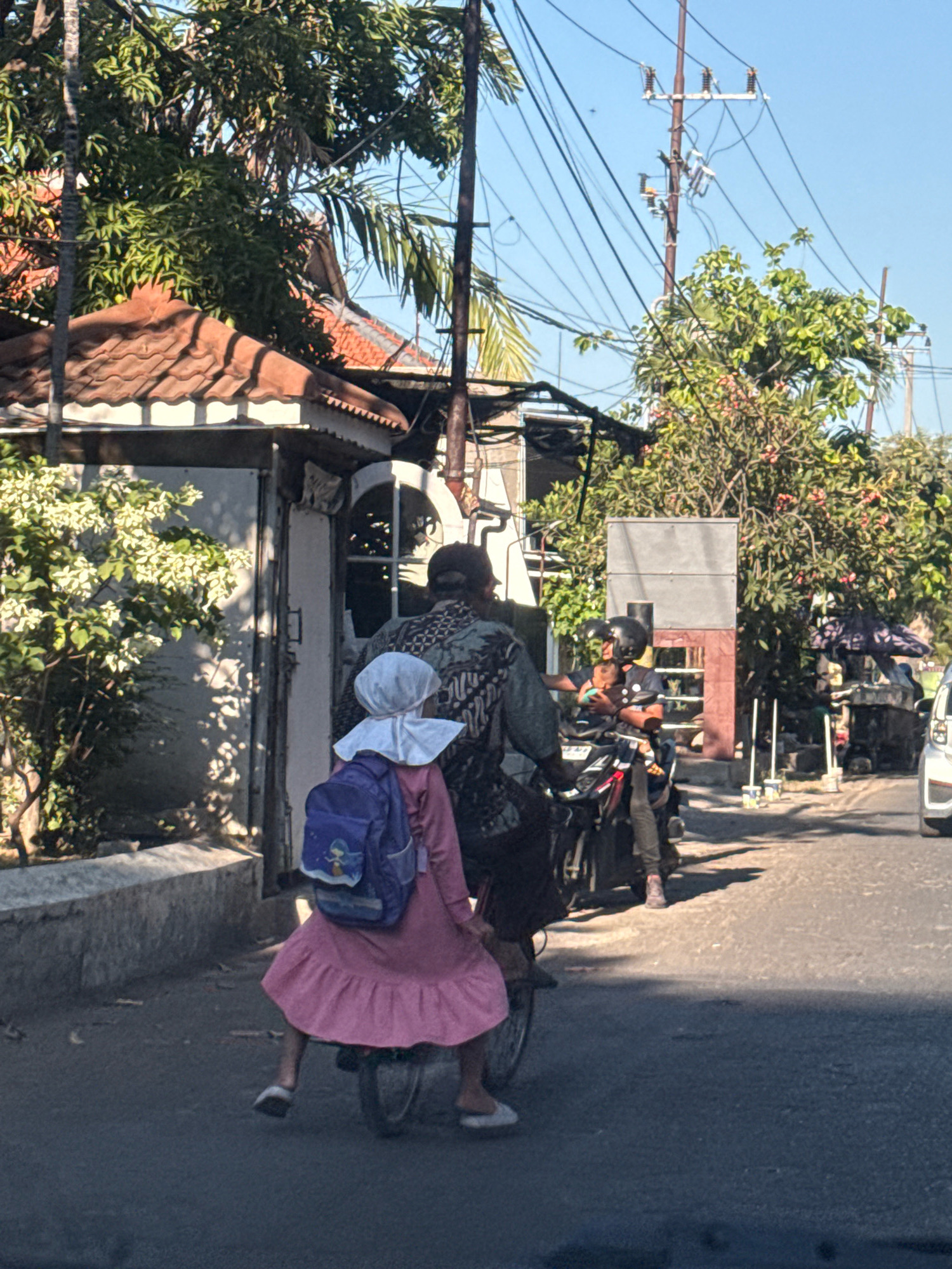 The image depicts a street scene in Surabaya, Indonesia, on a sunny day. In the foreground, a young child wearing a pink dress and a white headscarf is seated on the back of a motorcycle, with a blue backpack on their shoulders. The child's legs are visible, and they are wearing white shoes. The motorcycle is driven by an adult wearing a patterned shirt. In the background, another motorcycle is visible with two people on it, one of whom is holding a baby. Trees and overhead power lines are prominent elements of the environment, with buildings and tiled roofs also present. The road surface is asphalt, and there is a stone wall on the left side of the image. The overall scene suggests a typical mode of transportation in the city.