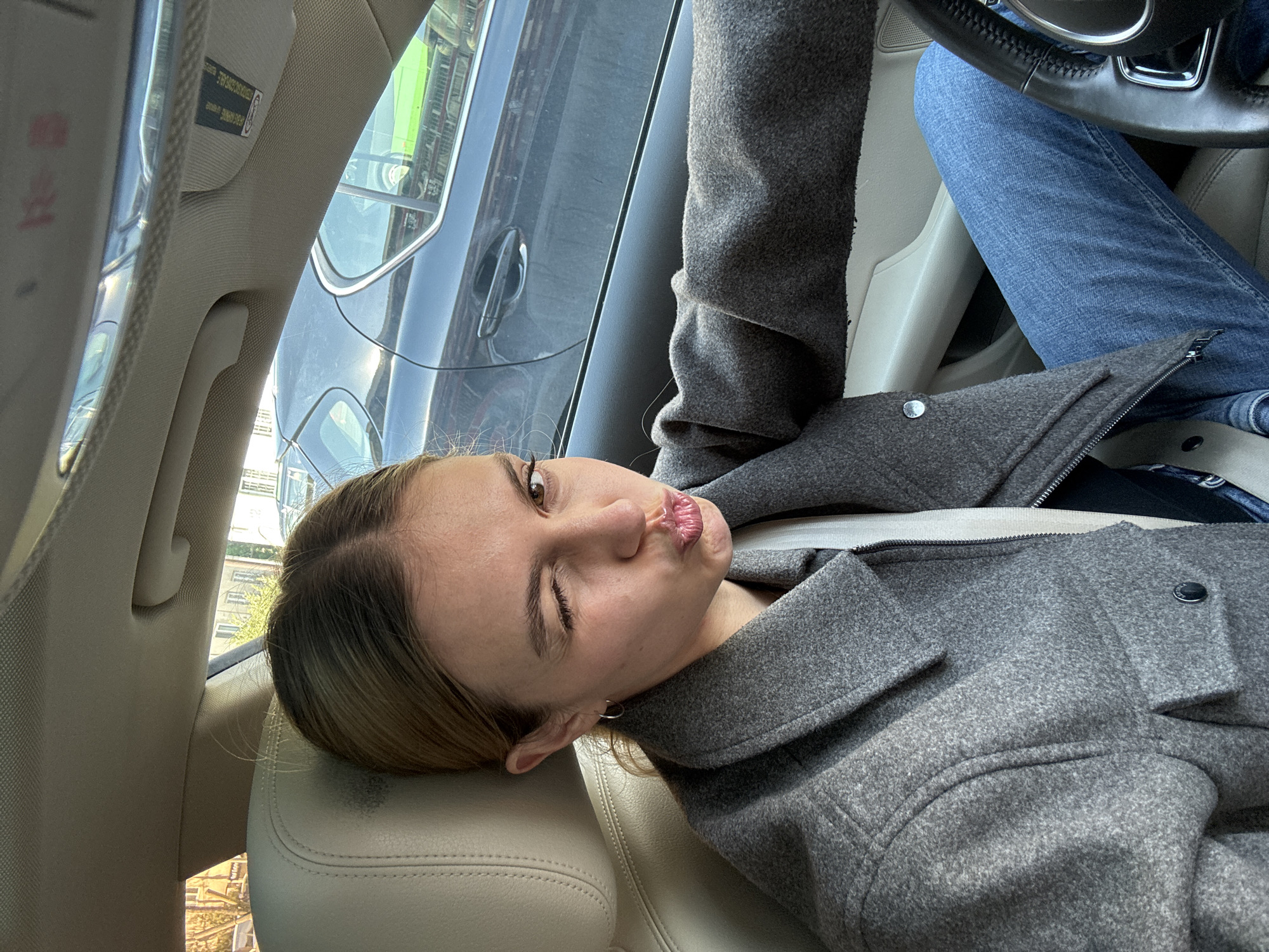 This media file features a tilted, casual portrait of a young woman inside a car, likely a selfie. She has light brown hair with blonde highlights and is wearing a textured grey coat and blue jeans. Her head rests on a light-colored car seat headrest. She makes a playful "duck face" or kissy face with pursed lips, winking with her left eye while her right eye is open, looking at the camera. A small silver hoop earring is visible in her left ear.
The car's interior is modern, with light-colored seats and a partial view of a black steering wheel with chrome accents. Outside the car, through the windows, an urban setting is visible, showing reflections of buildings, other vehicles (including a dark grey car with a door handle), and some greenery, consistent with a city environment like Tbilisi, Georgia. Natural daylight illuminates the scene, indicating it is daytime. A warning label, partially obscured and reflected, is visible on the sun visor. The image conveys a lighthearted and casual mood.