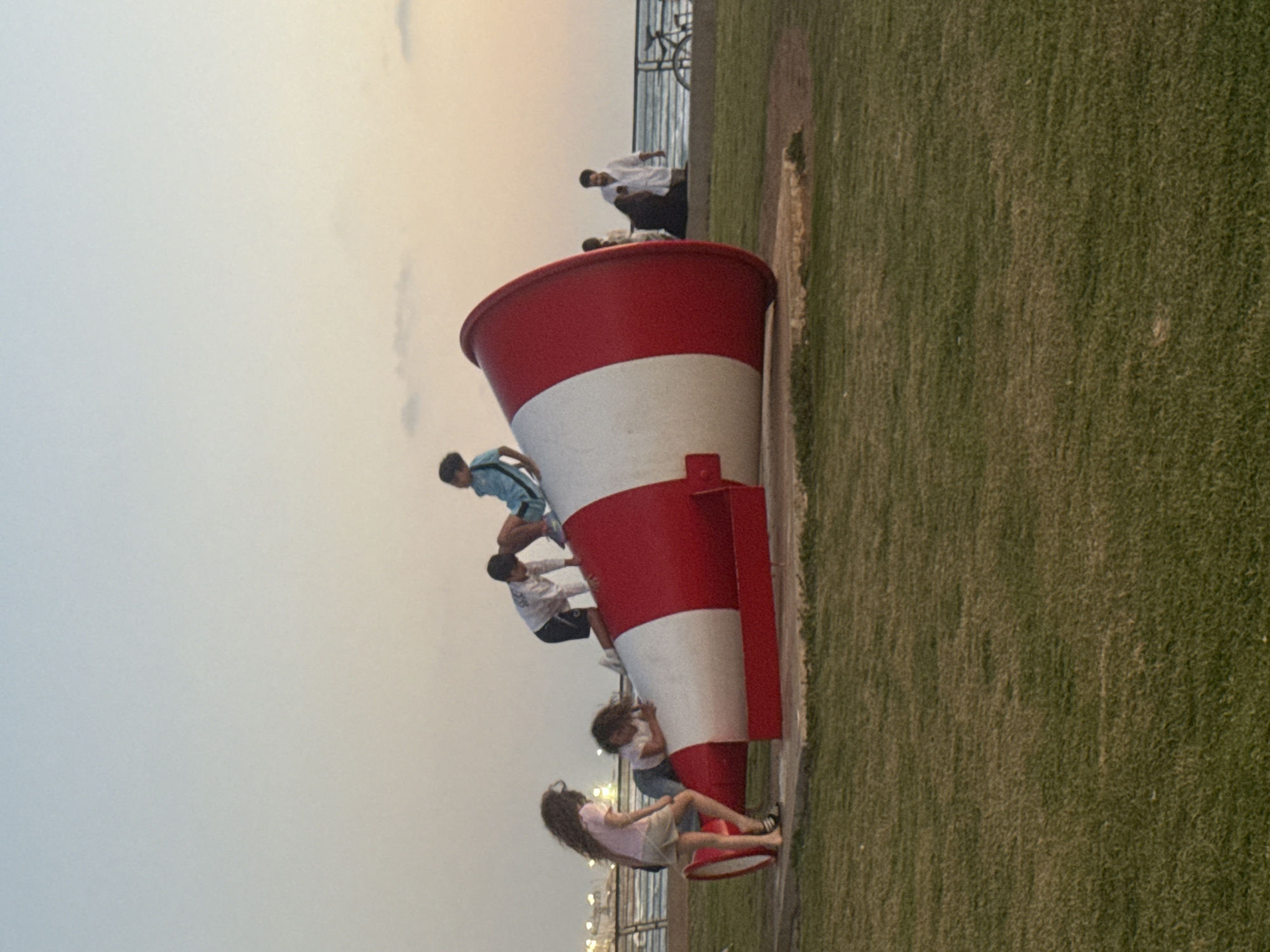 The image shows a large, red and white striped megaphone-shaped structure lying on its side in a grassy area. A group of people, including children, are climbing on the megaphone. The background shows a fence, a walkway, and blurred figures of people standing near the water. The sky is overcast. The location is Tel Aviv, Israel.