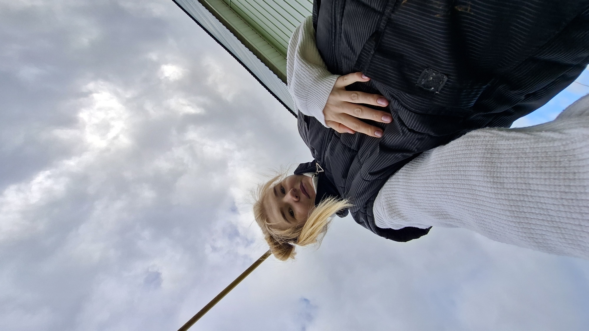 The image shows a woman standing outdoors in Большой Букор, Russia. The perspective is from a low angle, looking up at the woman. She is wearing a white sweater and a black puffer vest. Her blonde hair is styled in small buns. Her left hand is visible near her chest. Overhead, there's a cloudy sky. A thin pole is visible in the lower left corner of the image. The edge of a green roof is visible at the very top.