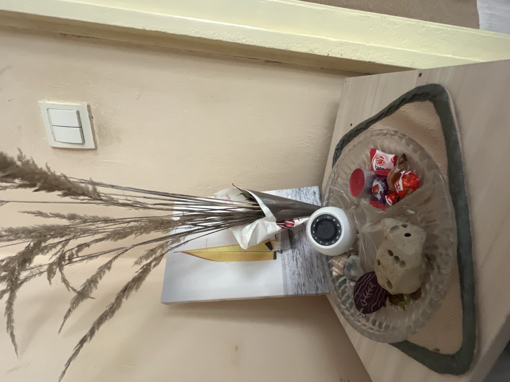 A slightly rotated high-angle shot captures a still life arrangement on a surface, likely a table or shelf, situated against a light beige wall. On the wall, a standard double light switch is visible. The arrangement includes a transparent glass dish with a braided rope rim, containing assorted small items such as candies, shells, a small rock with holes, and a decorative wooden piece. Next to the dish is a white, spherical device, possibly a small security camera or a similar electronic device. A print featuring a yellow sailboat against a seascape provides a backdrop to a bunch of dried wheat stalks, adding a natural element to the composition. The location is unknown, however, based on the metadata it could be Varna, Bulgaria.
