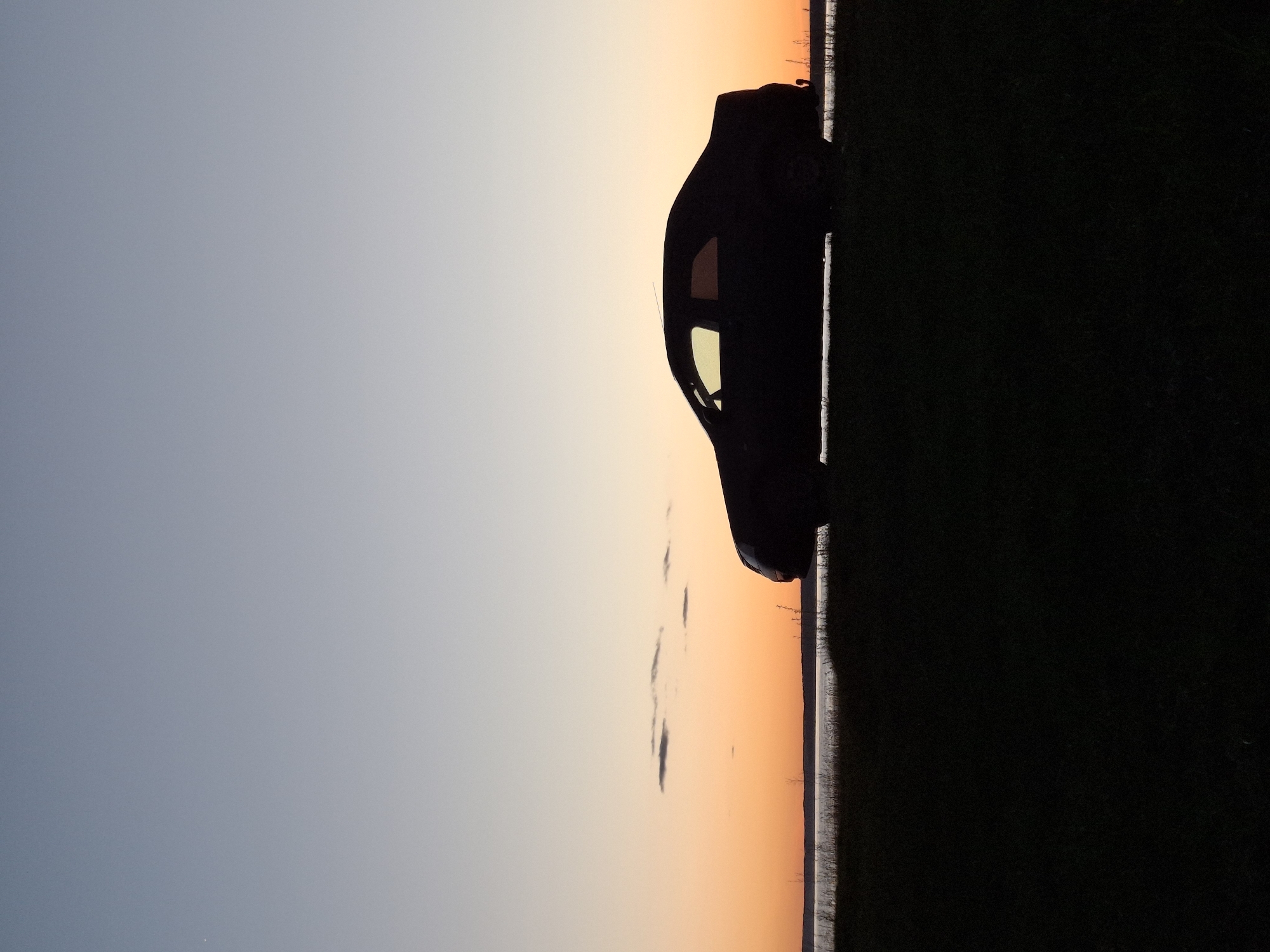 The image captures the silhouette of a car against a backdrop of a gradient sky transitioning from light gray-blue to orange, suggestive of a sunset or sunrise. The car is parked on a strip of land adjacent to a dark, undefined area, likely foliage. The location is Plishkino, Russia. No people are visible.