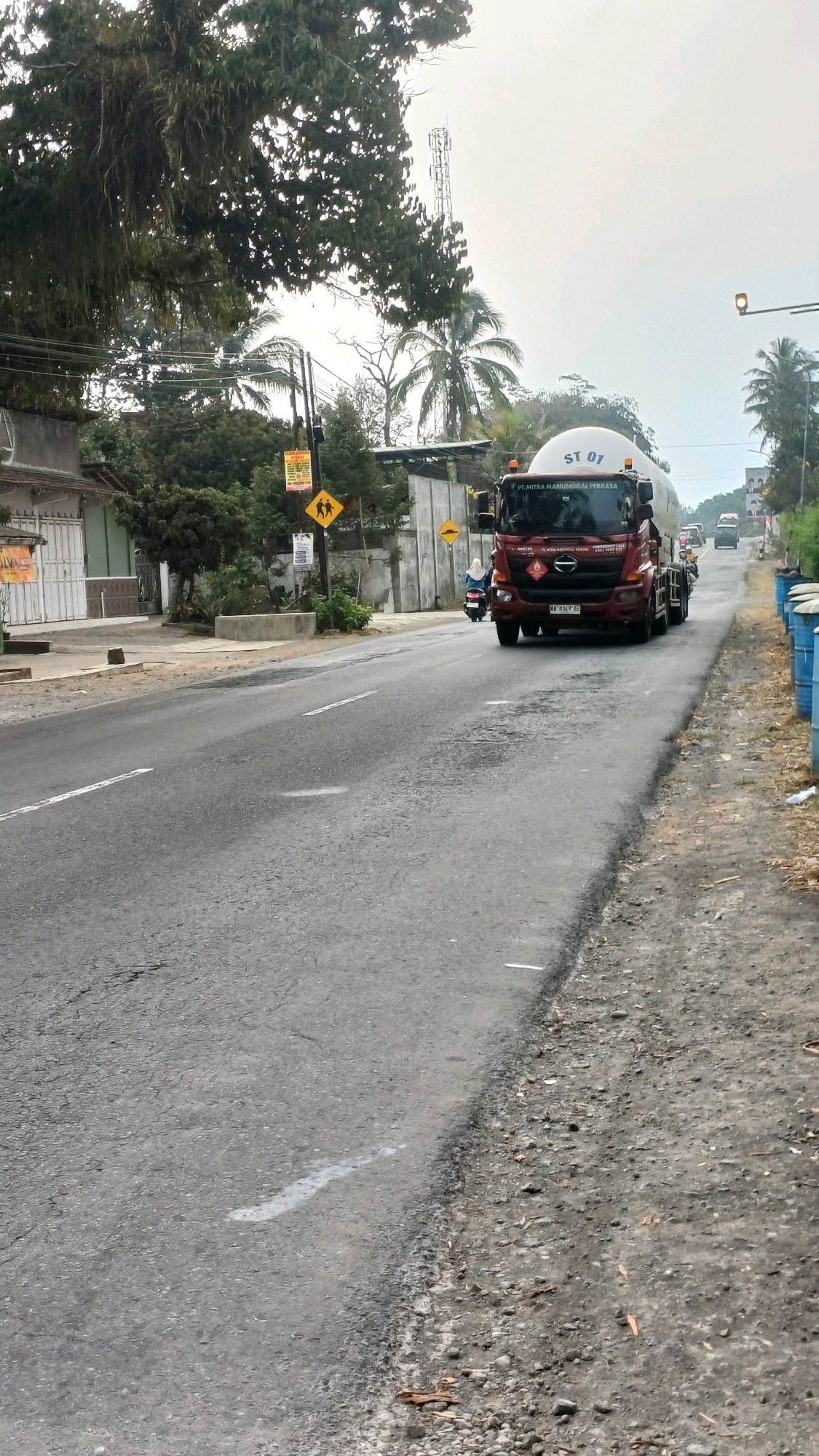 The image depicts a road scene in Tengahan, Indonesia, under an overcast sky. A red fuel tanker truck, identified with "ST 01" and "PT. MITRA MANUNGGAL PERKASA" markings, is traveling on the asphalt road. A scooter with a rider is in front of the truck. The road is lined with trees on the left and right, with buildings and a cell tower visible in the background. Several blue barrels are stacked along the right side of the road. Traffic signs, including one depicting two figures walking, are also present. The road surface shows signs of wear and has white lane markings.