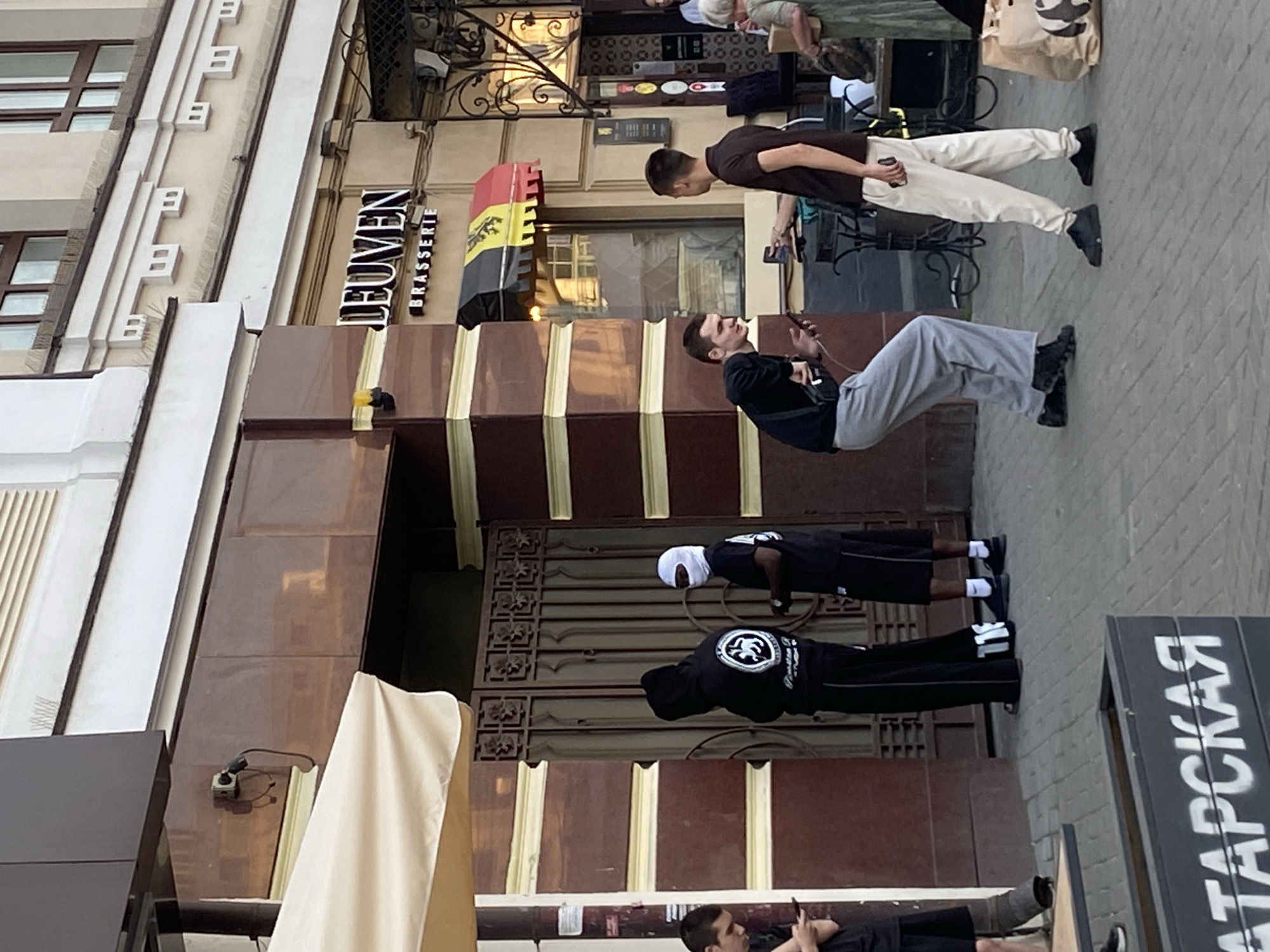 The image depicts a street scene in Kazan, Russia. A building with the sign "LEUVEN BRASSERIE" is visible, indicating the presence of a restaurant or brewery. Several people are present in the scene. Two individuals are standing on the sidewalk, appearing to be tourists or locals observing the surroundings. Another individual is leaning against a wall, seemingly occupied with their mobile phone. Two individuals are engaged in push-ups in front of the restaurant's entrance. The environment includes a building facade with detailed architectural elements, a sidewalk, and signage in Russian on a black rectangle. Overall, the image captures a blend of urban activity and street life in Kazan.