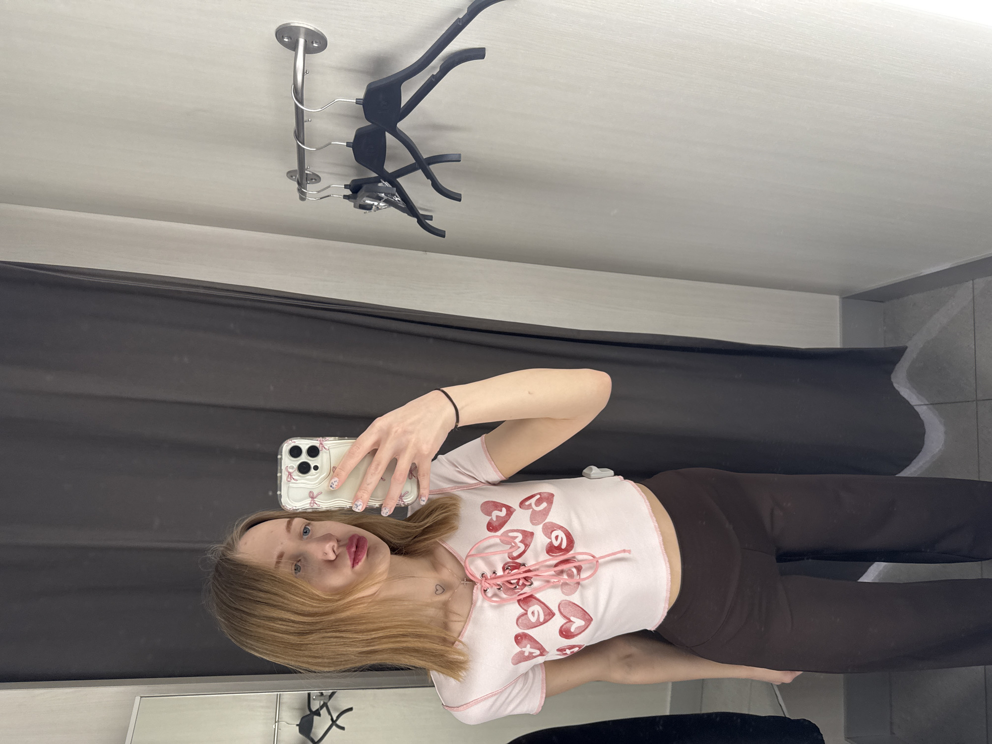 The scene depicts a young female inside a changing room in a retail establishment in the city of Ovoshchnoy, Russia. The female, with shoulder-length blonde hair and blue eyes, has a small stud piercing on her left nostril and is wearing red lipstick. She is attired in a light pink, short-sleeved crop top featuring a laced V-neck closure and multiple red heart graphics with white numbers, paired with dark brown loose-fitting pants. A silver necklace with a heart pendant and a cross pendant is visible around her neck, and a black elastic band is on her right wrist. Her fingernails are manicured with light polish and small decorative elements. She is holding a white smartphone, fitted with a clear case decorated with pink bows, in her right hand, positioned as if taking a self-portrait. Her lips are slightly pouted. The changing room features light grey textured walls. A dark grey or black privacy curtain is visible on the left. A stainless steel hanging bar with three black plastic clothing hangers is mounted on the right wall; one hanger has a security tag attached. The floor is tiled light grey with a darker grey baseboard.