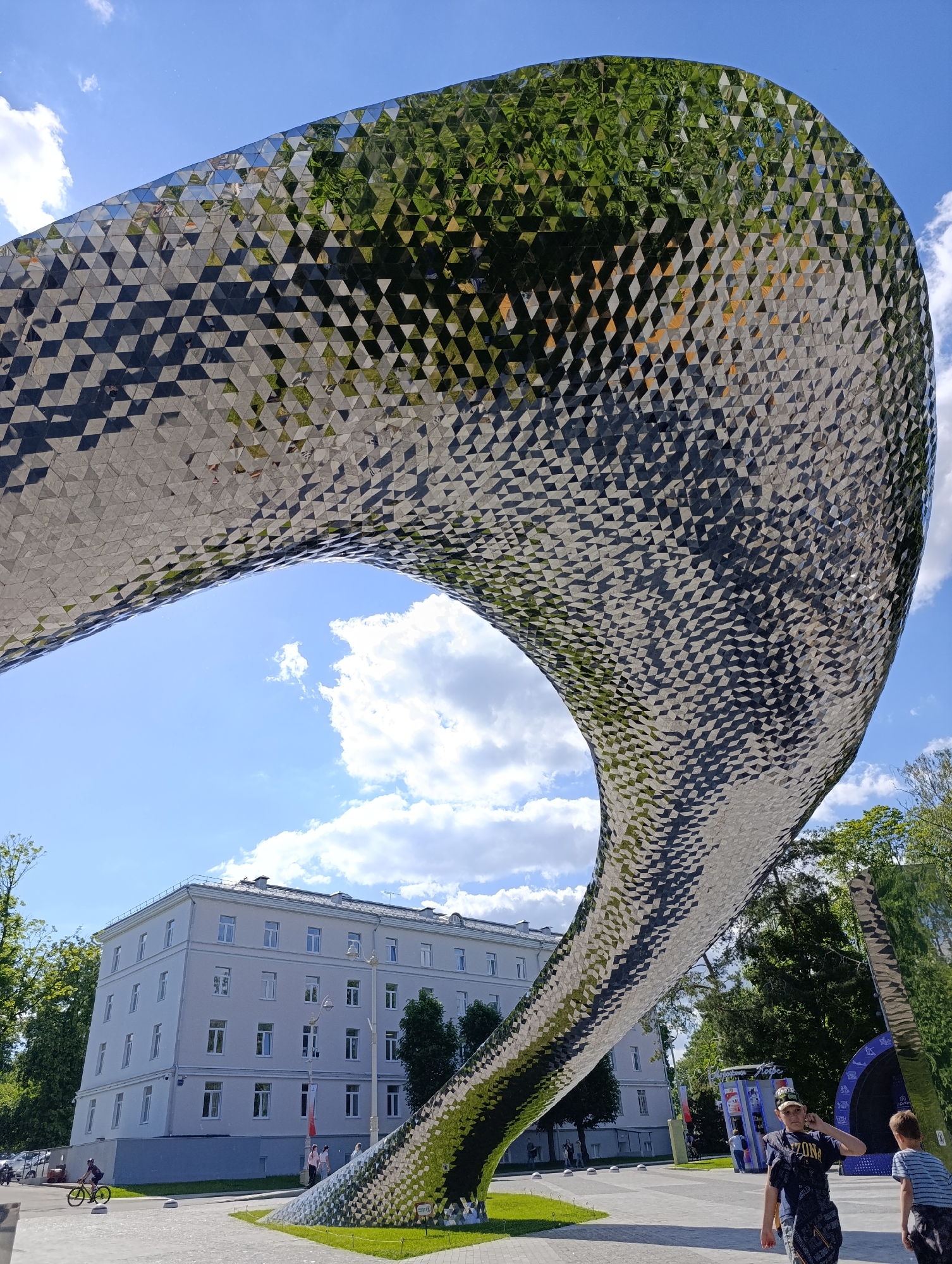 The image features a large, flowing, and highly reflective sculpture, appearing to be made of numerous small, mirrored triangular facets. The sculpture curves significantly, forming an arch-like structure that frames a light-colored, multi-story building with multiple windows in the background. The surface of the sculpture vividly reflects the bright blue sky, scattered white clouds, and lush green trees that surround it.
The scene is an outdoor public space, likely a park or square, on a sunny day. In the foreground, two boys are visible, one wearing a dark shirt with "ARIZONA" text and a cap with partially visible text like "ZONE A". They are standing on a paved area next to a patch of grass. Further back, a person is seen riding a yellow bicycle, and other individuals are walking near the building. The ground is a mix of paved pathways and green lawns, with some bollards lining the edge of the path. A streetlamp stands near the building. Small, indistinct text or symbols are visible on signs attached to the base of the sculpture. A red and white banner, with unreadable text, hangs from the building.
The overall impression is a modern urban landscape in Moscow, Russia, where art and architecture blend with natural elements under a clear, bright sky.