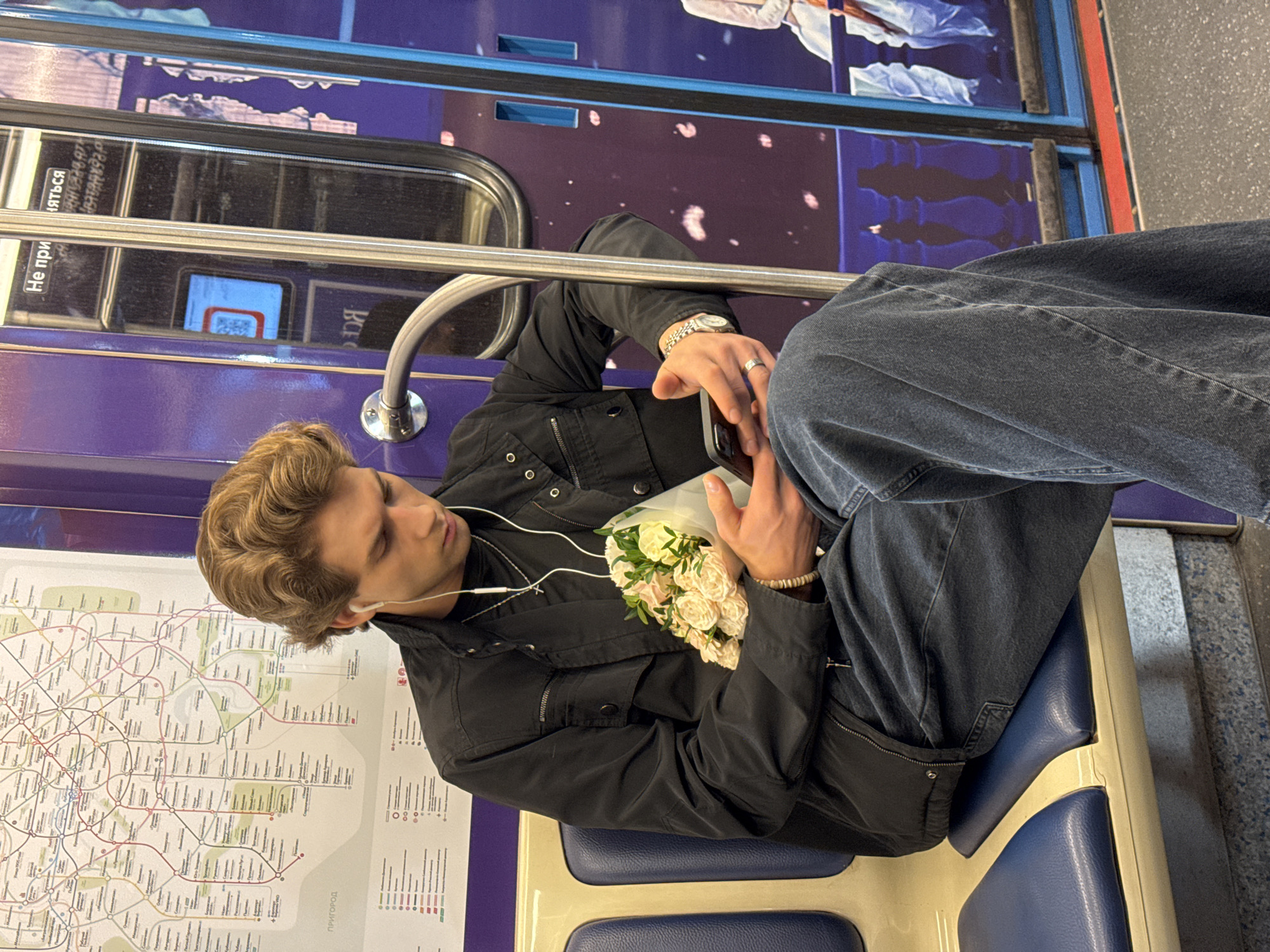 A young man is sitting on a train in Moscow, Russia. He is holding a bouquet of flowers and looking at his phone. He is wearing a black jacket, jeans, and has headphones in. The train car has a purple interior with a map of the Moscow Metro system on the wall.