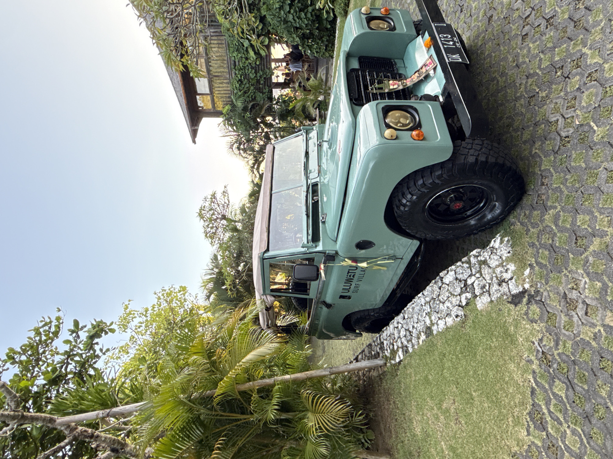 The image, rotated 90 degrees counter-clockwise, displays a light green vintage-style off-road vehicle, resembling a Land Rover Defender, parked in what appears to be a resort or villa complex in Kuta Selatan, Indonesia. The vehicle is equipped with rugged tires and features a soft top. Adorning its side is the text "ULUWA**T**U SURF VILLAS", identifying its affiliation with a local establishment. The license plate, "DK 1413 T", is clearly visible on the front bumper, indicating a registration from Bali, Indonesia. Decorative elements, possibly traditional Balinese offerings or flowers, hang from the front grille and side mirror.
The vehicle rests on a patterned paved surface interspersed with grass, adjacent to a low stone wall and a lush green lawn. The setting is rich with tropical foliage, including prominent palm trees and other broad-leafed plants, suggesting a warm, humid climate. In the background, a multi-story wooden building with large windows, typical of resort architecture, is partially obscured by dense greenery. Blurry figures of people can be vaguely discerned inside the building in one of the crop images, suggesting activity there. The clear, bright sky and soft lighting indicate either early morning or late afternoon. The overall scene conveys a serene and tropical environment, likely a holiday destination.