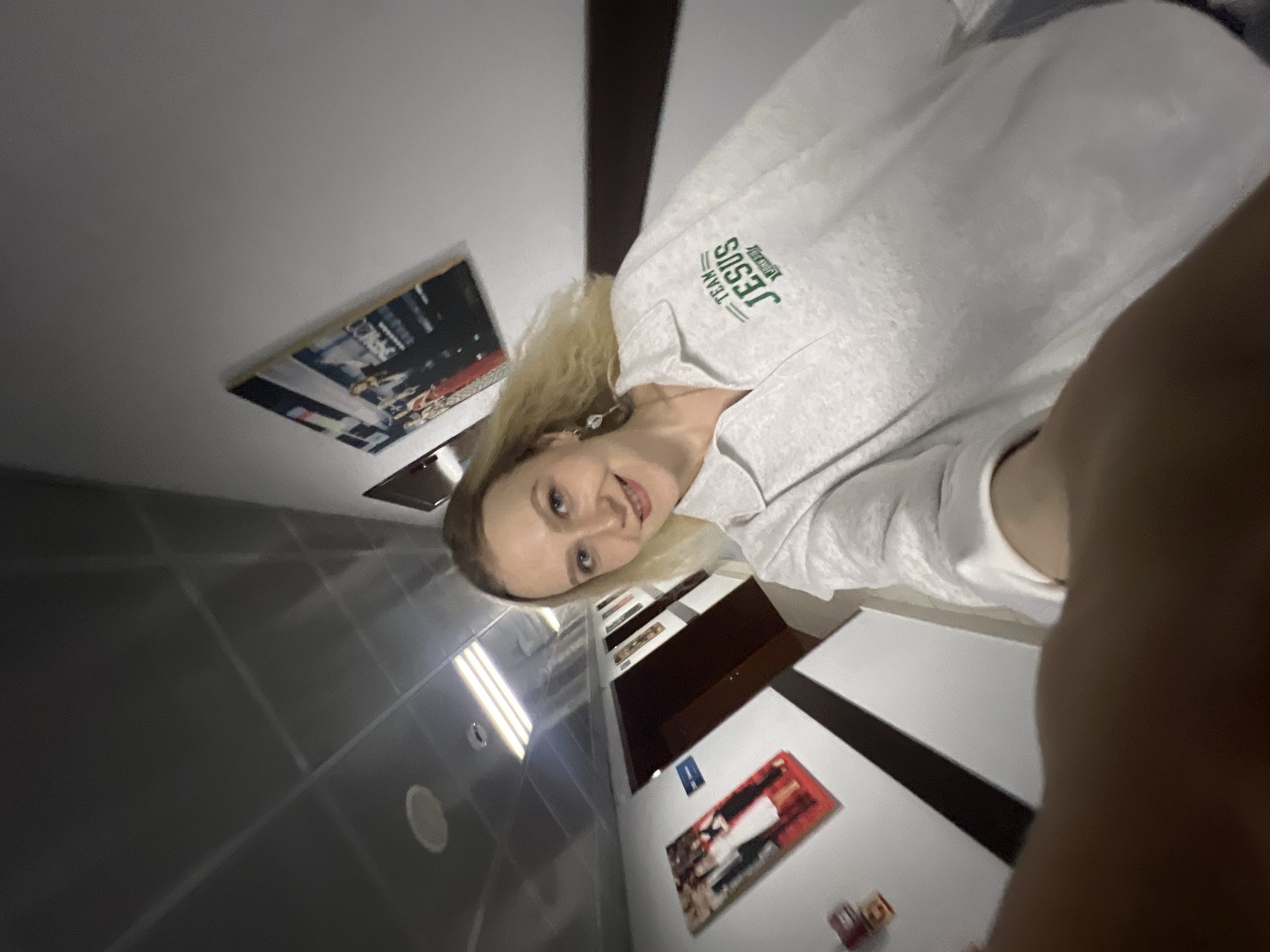 A blonde woman with blue eyes is taking a selfie in a hallway. She is wearing a light gray sweatshirt with green lettering that reads "TEAM JESUS". The hallway appears to be indoors, with a drop ceiling and fluorescent lighting. There are framed pictures on the walls, including what appears to be a wedding photo and another group photo. A dark wooden door is visible to the left of the woman, and a sign with a blue symbol is posted on the wall next to it. The floor is tiled with dark and light sections. The woman is smiling and looking at the camera. The overall scene is well-lit and appears to be in a professional or institutional setting.