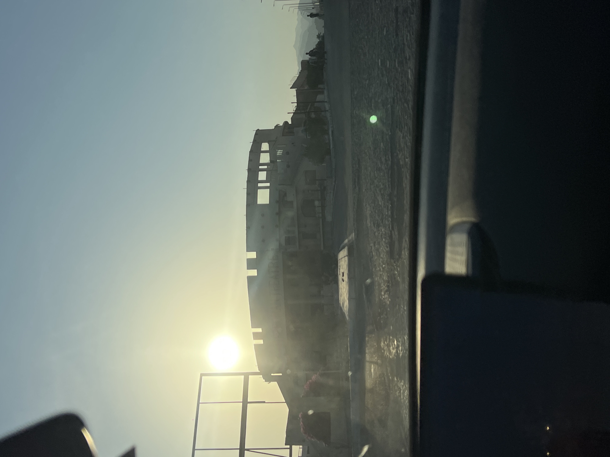 The image depicts a sun-drenched, open-air structure resembling a coliseum or amphitheater, located in Sharm El Sheikh, Egypt. The structure, made of what appears to be concrete or stone, has an unfinished, weathered appearance. The sun is high in the sky, casting a bright glare and creating silhouettes of people walking in the distance. The environment appears arid and mountainous, typical of the Sinai Peninsula. The foreground shows a portion of a car's interior, suggesting the photo was taken from a moving vehicle. The overall scene evokes a sense of a historical site or a construction project in a desert landscape.