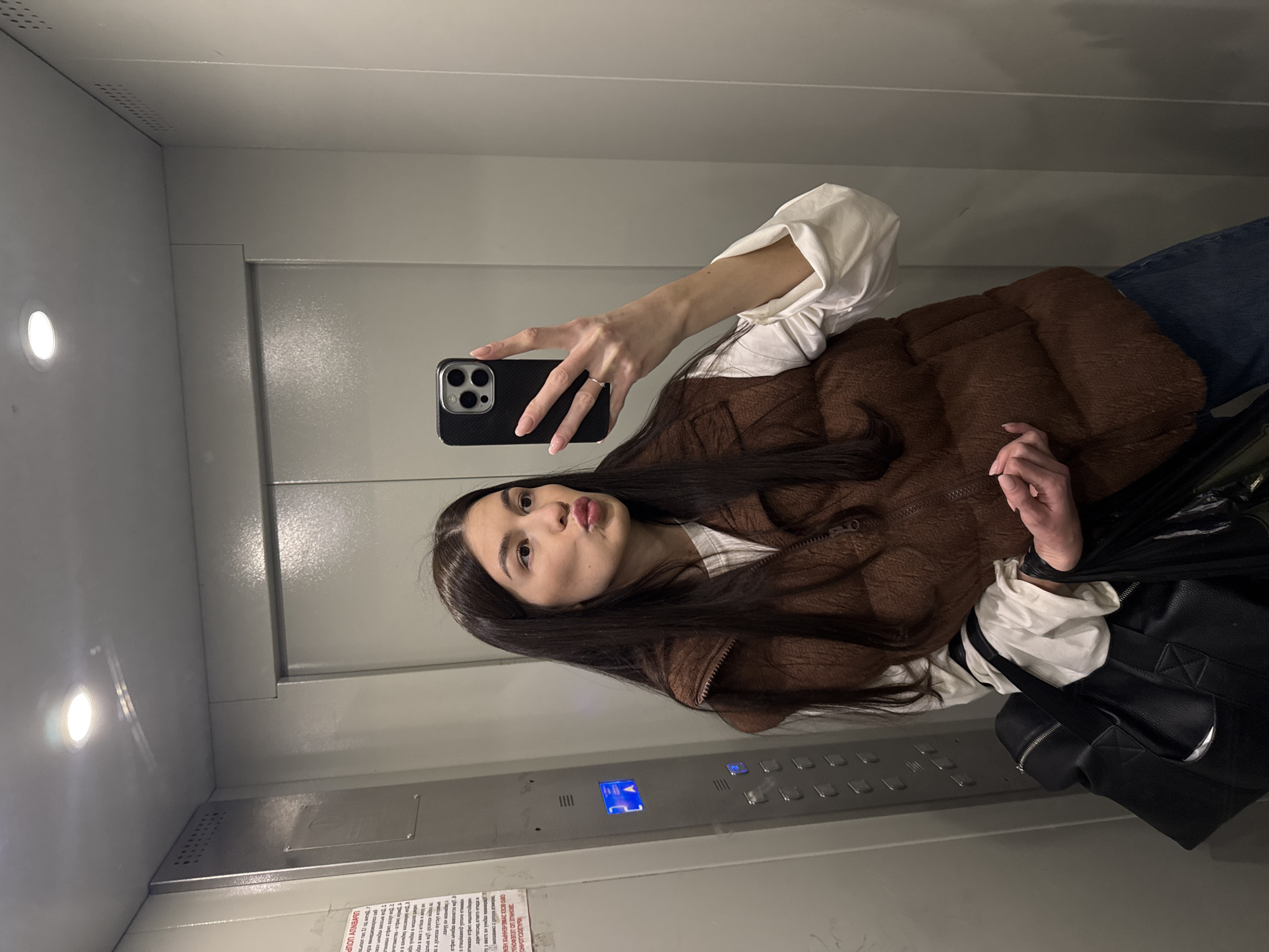 A young woman with long brown hair is taking a selfie in an elevator. She is wearing a white shirt with rolled-up sleeves and a brown vest. She is holding a black iPhone with a triple-lens camera, and her expression is a slight pout.
The elevator's interior is metallic gray. Control buttons are visible on the wall, along with a printed notice in Russian. A black purse is partially visible to the right of the woman, suggesting she may have other belongings with her. The image was taken in the city of Chaikovsky, Russia.