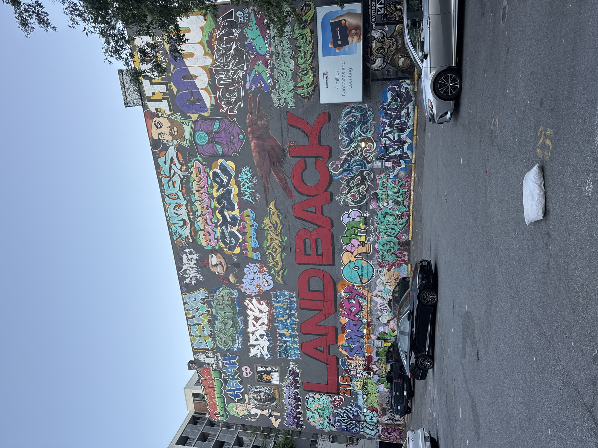 The image displays a large, colorful mural covering the side of a building, likely in an urban setting. The mural features a variety of graffiti art, including large, bold lettering that spells out "LAND BACK" in red. Other elements of the mural include stylized portraits, abstract designs, and depictions of animals, such as a prominent red bird.
Below the mural, a parking lot is visible with several cars parked. A black sedan is in the foreground, and a silver sedan is parked to the right. A black pickup truck is parked further back. The ground of the parking lot is asphalt, with some yellow markings and occasional debris.
In the background, the upper floors of adjacent buildings are visible, some with balconies. A billboard for Capital One is mounted on the wall of the building with the mural. Trees with green foliage are present on the right side of the image, partially obscuring the top of the building. The sky above is clear and blue, indicating daytime.
This mural is located in Vancouver, Canada, specifically in the downtown area. This particular mural is known to be situated on the south-facing wall of the historic Orpheum Theatre, a renowned performing arts venue. The "LAND BACK" message is a significant political statement, advocating for the return of Indigenous lands to their original stewards. The artwork on the wall is a dynamic and layered composition, with various styles and eras of graffiti art coexisting.
