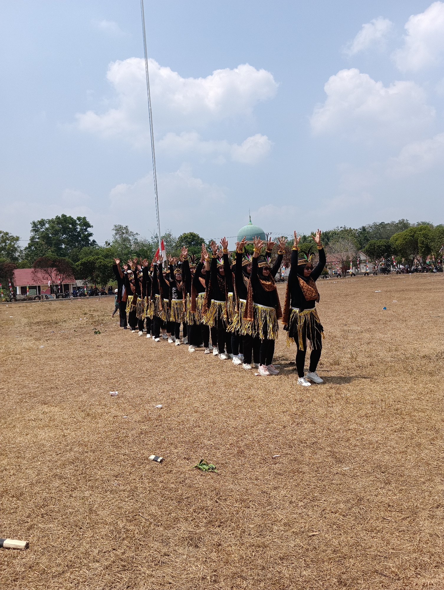 In the city of Pematang Jaya, Indonesia, a group of individuals, primarily women, are participating in a cultural performance on a dry, grassy field. They are lined up in a diagonal formation, facing towards the right of the frame, with their arms raised in unison. The performers are dressed in traditional black attire, adorned with decorative gold fringe and headdresses. Some wear sunglasses, and all are wearing white athletic shoes.
The background reveals a building with a green dome, likely a mosque or religious structure, situated amidst trees. In the distance, there are more trees, a red-roofed building, and various vehicles, including ATVs, suggesting a public event or gathering. The sky is partly cloudy, with a tall flagpole visible to the left of the performers. The ground is covered in dry, straw-like grass, with a few scattered white paper scraps and a green rope visible. The overall scene depicts an outdoor cultural event under a bright, daytime sky.