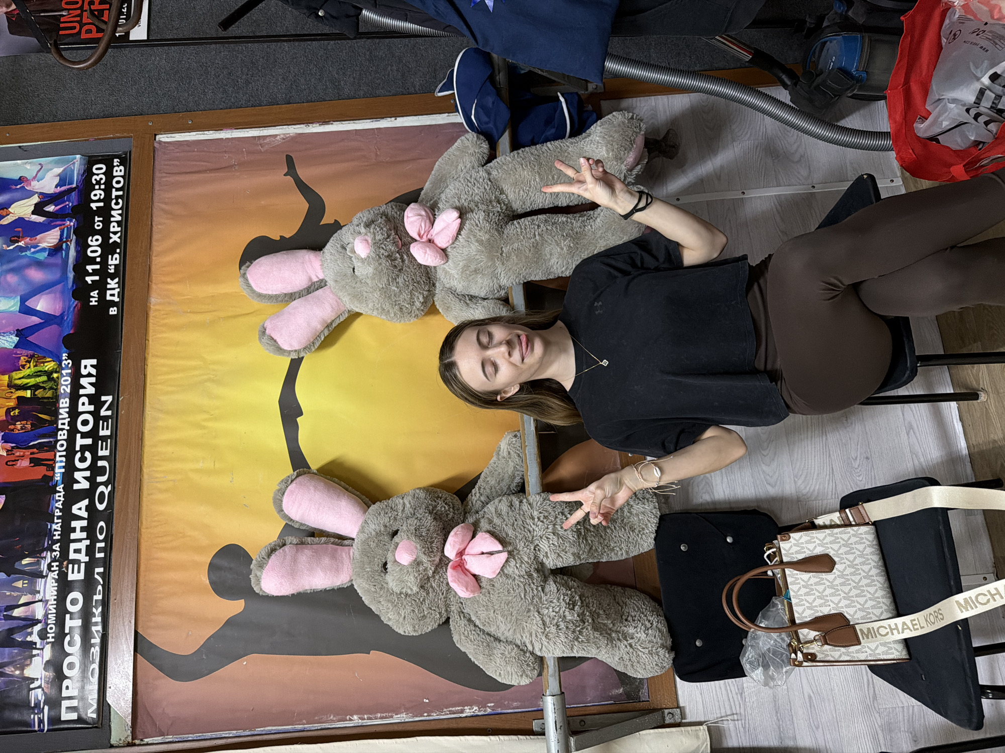 A woman is sitting in a chair in what appears to be a storage room, with two large, gray plush bunny toys beside her. She is wearing a black t-shirt and brown leggings, and she is making a peace sign with both hands. Beside her chair is a black shopping bag and a white Michael Kors handbag. In the background is a poster for a musical called "Simply a Story" that is based on the music of Queen, and which was nominated for a "Plovdiv 2013" award. It lists the performance location as "DK 'B. Hristov'" on June 11 at 7:30 PM. The poster includes an image of dancers. There is also a vacuum cleaner and other storage items in the background. The location is possibly in Plovdiv, Bulgaria, based on the text on the poster.