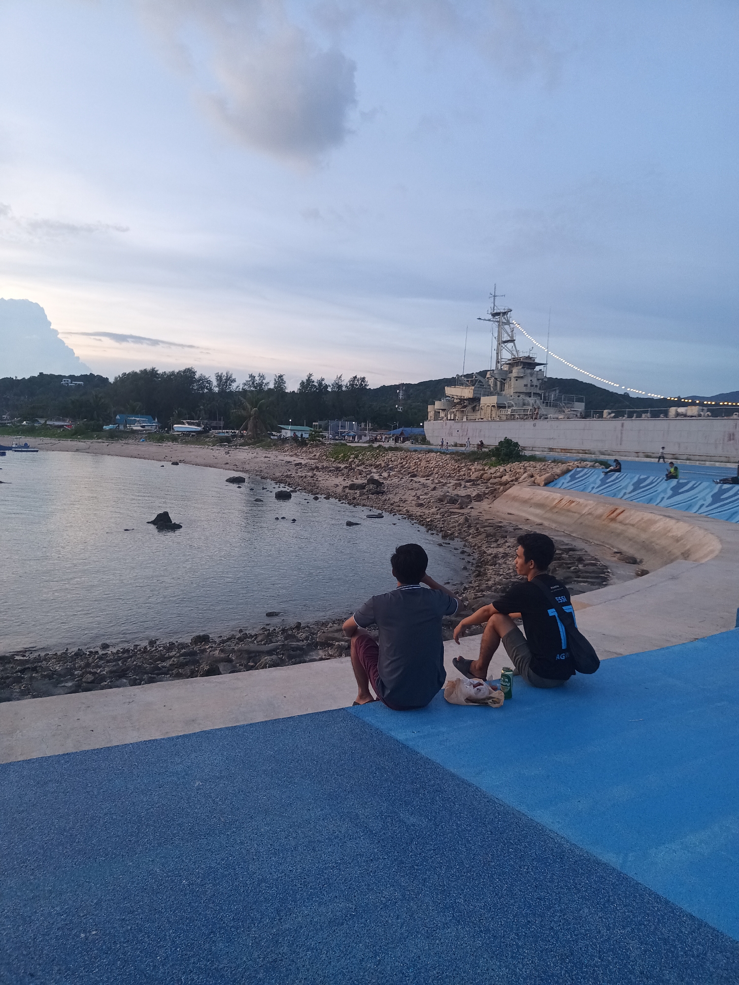 The image depicts a serene waterfront scene at dusk in Koh Phangan, Thailand. Two individuals are seated on a blue, textured pathway, facing away from the camera and towards the water. The person on the left is wearing a dark grey t-shirt and maroon shorts, while the person on the right is in a black t-shirt with a prominent number '7' and grey shorts. Next to them on the pathway are a paper bag and two cans, suggesting they are having a relaxed break.
The pathway is elevated and curves along the coastline, with a rocky shore bordering the calm water. In the background, a large, light-colored ship is docked, its structure illuminated by strings of lights. Beyond the ship, a densely forested hillside rises, with some buildings visible among the trees. The sky above is a soft gradient of blue and grey, with scattered clouds, indicating the transition from day to night. The overall atmosphere is peaceful and contemplative.