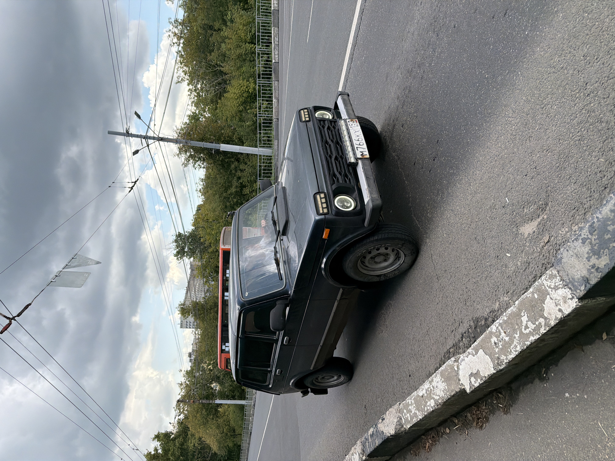 The image captures a dark-colored Lada Niva SUV driving on a paved road in Nizhny Novgorod, Russia. The vehicle is positioned in the right lane, with its front facing slightly to the left. A person is visible inside the SUV, in the driver's seat. The road is bordered by a concrete curb on the left, which shows signs of wear and tear. In the background, a red bus is partially visible on a road ahead. Trees line the distant background, with buildings also visible beyond the trees. Overhead, power lines and a street light pole are prominent. The sky is overcast with dramatic clouds. The license plate on the Lada Niva reads "M766KX 152".