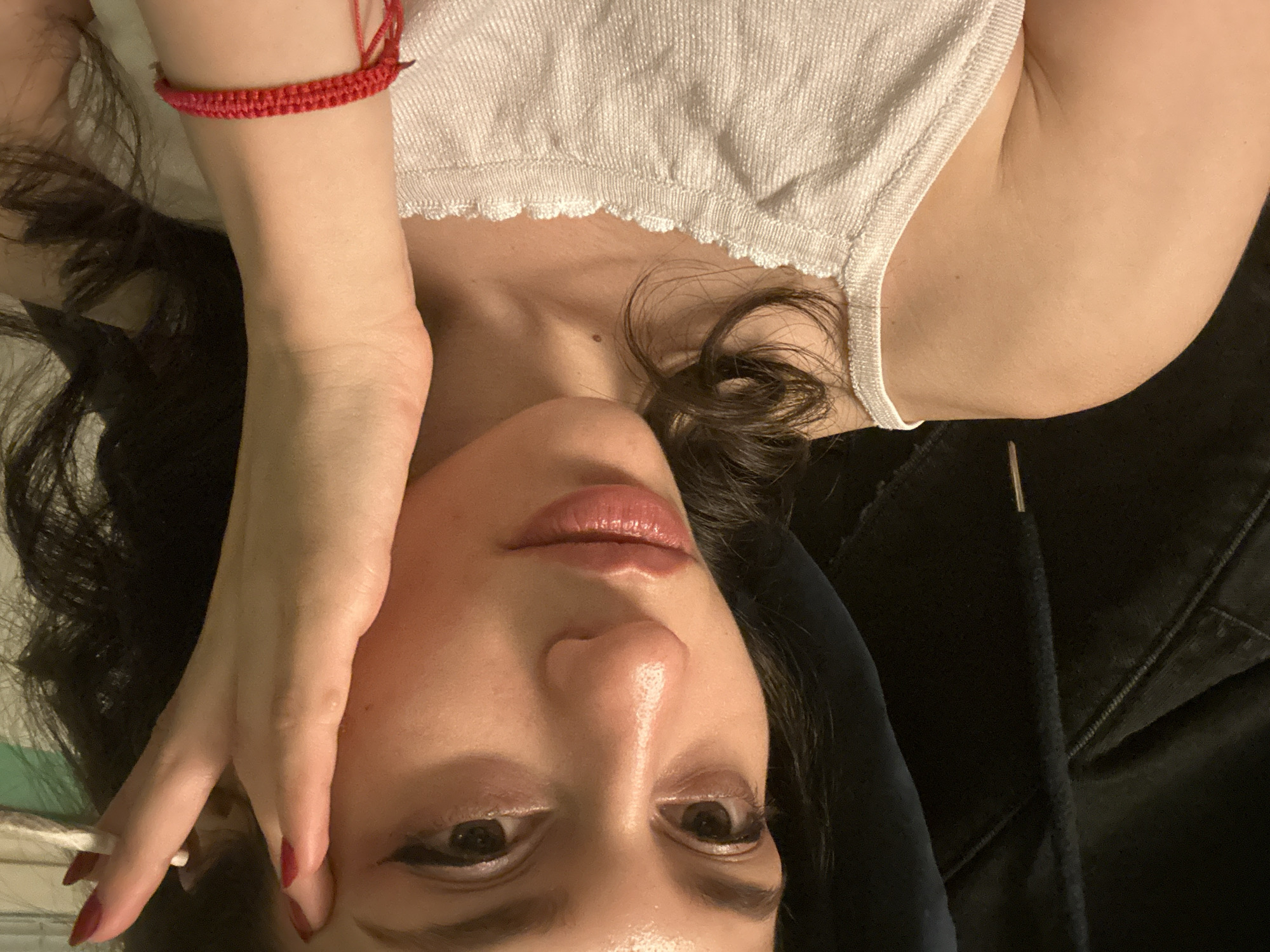A woman with dark hair and red nail polish is shown in a close-up shot, facing the camera. She is wearing a white tank top and a red bracelet. Her right hand is raised to her forehead. The background is dark and appears to be indoors. There is no identifiable information that can place this image in Sofia, Bulgaria.