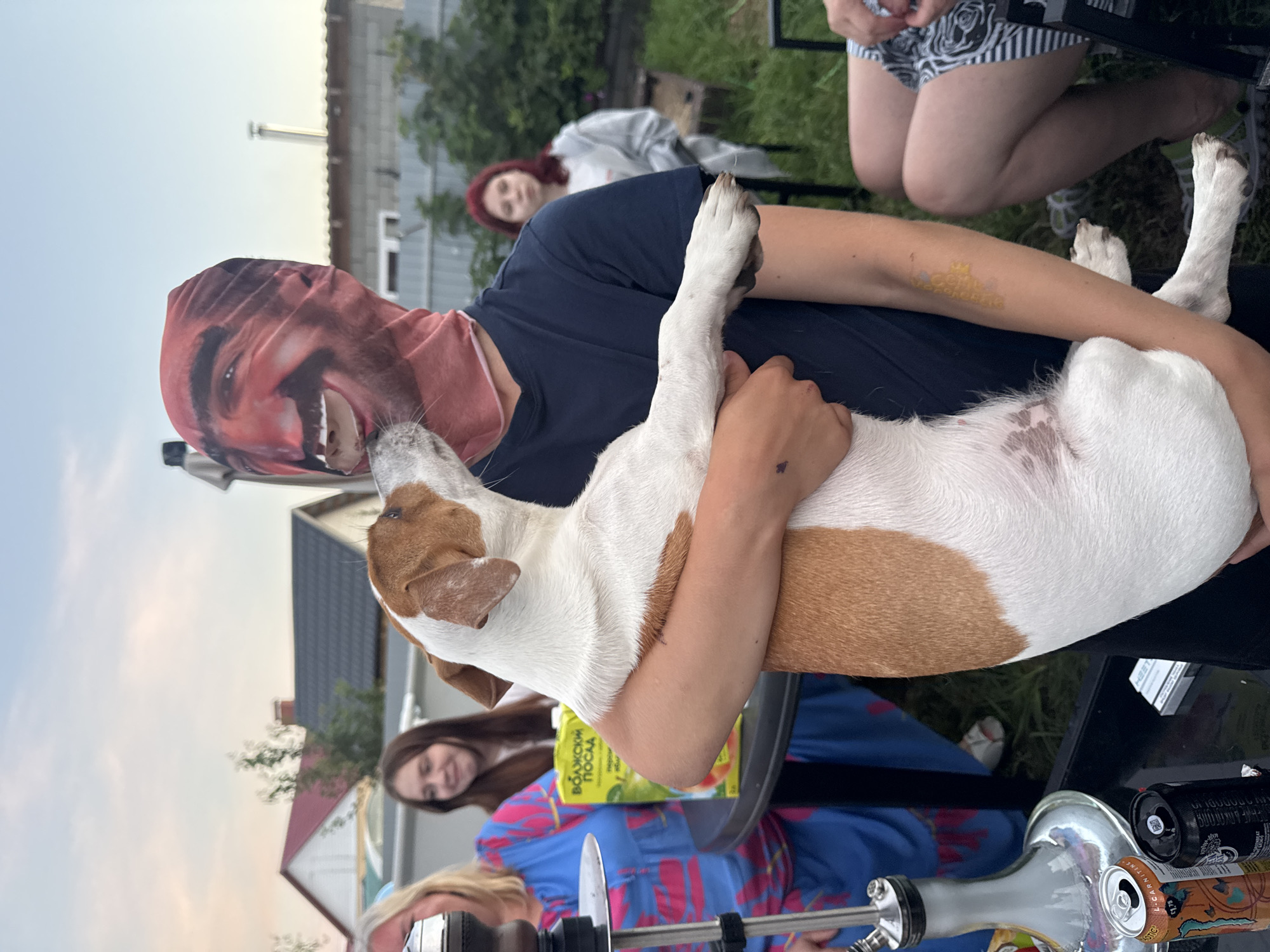 In Узюково, Russia, a person wearing a full head mask featuring a printed face is holding a Jack Russell Terrier dog. The dog is licking the mask on the person's mouth area. In the background, several people are visible. To the left, a woman in a blue dress with pink floral patterns is standing near a hookah. Another woman with dark hair is standing further back, smiling. To the right, a woman with red hair is looking away from the camera. The scene appears to be an outdoor gathering, possibly a backyard or garden, with trees and houses visible in the background. There are tables with drinks and other items on them, including cans and a juice carton. The sky is partly cloudy.