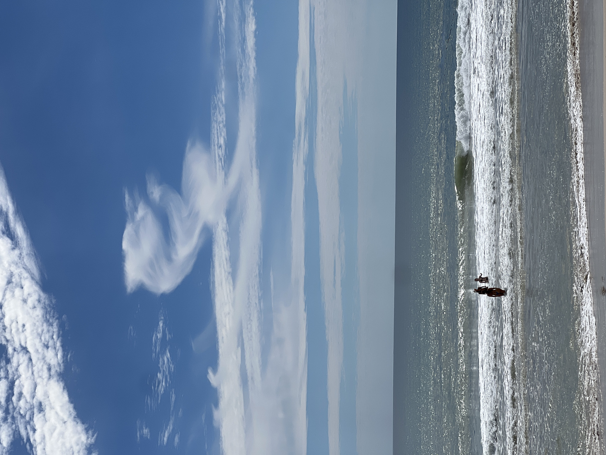 A bright, sunny day at an unknown beach with a clear blue sky and scattered clouds. The ocean is a deep blue with white-capped waves rolling onto the shore. Three people are in the water, two adults and one child, wading in the shallows. The foreground shows the sandy beach with a gentle wave washing ashore. The overall scene depicts a peaceful day at the beach, with people enjoying the water.