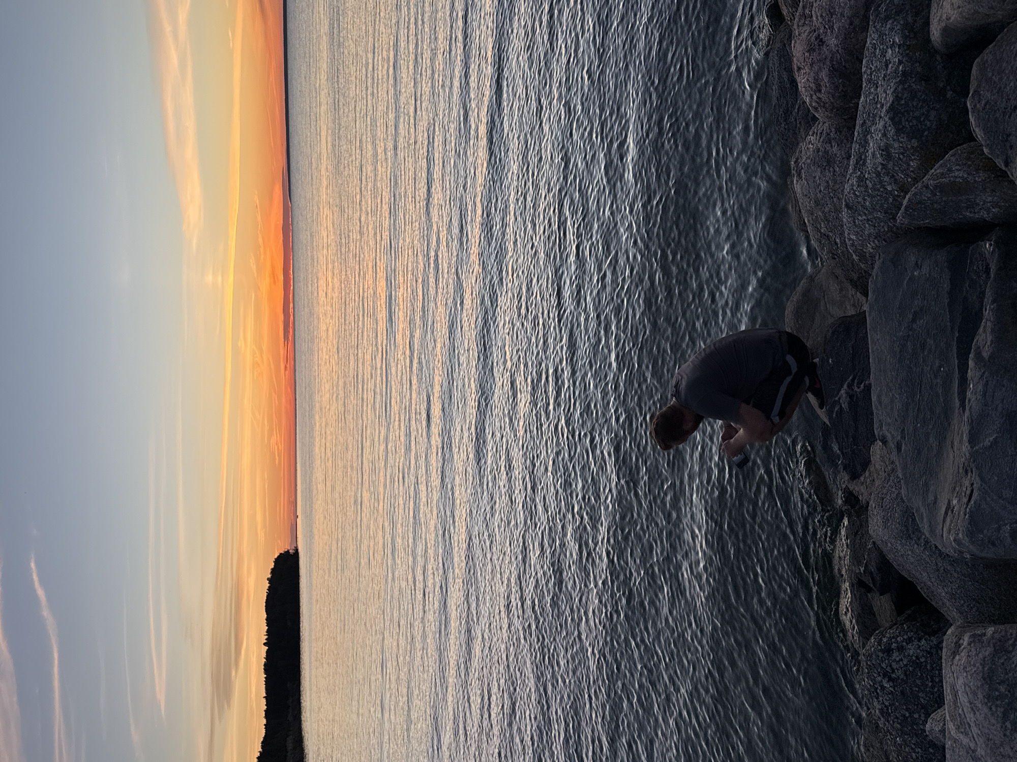 A full shot captures a sunset over the Baltic Sea, with a man in a grey t-shirt and black shorts squatting on a rocky shore. The man is holding a phone or camera in his hands, looking down at it. The water is calm with ripples reflecting the orange and pink hues of the sky. A dark tree line forms the distant horizon on the left, and the sky transitions from a light blue to a vibrant orange and pink as it meets the sea. The image appears to have been taken from a ground-level perspective, emphasizing the texture of the rocks and the vastness of the water and sky. The location is identified as Tallinn, Estonia.