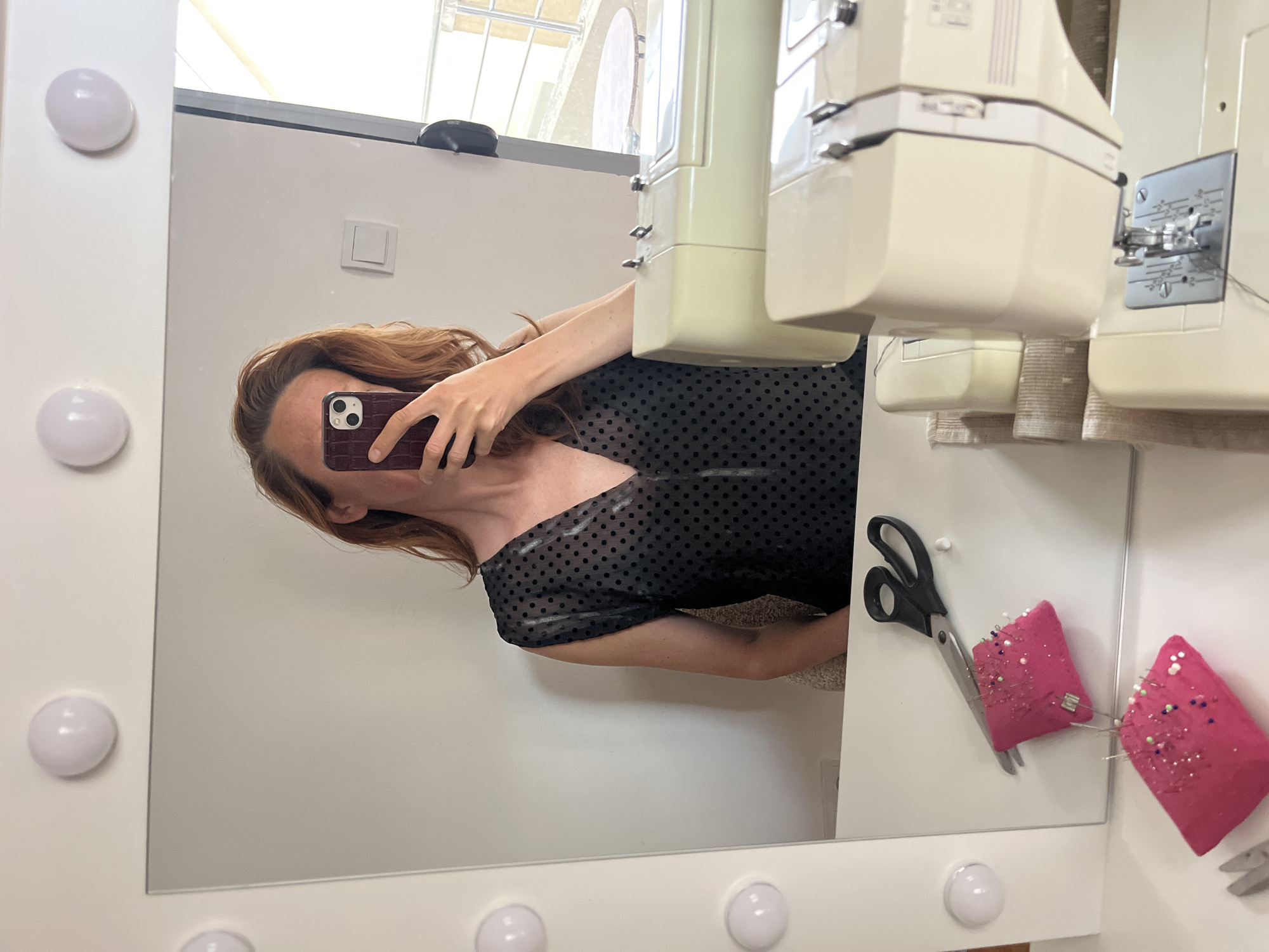 The image is a reflection in a vanity mirror with light bulbs around the perimeter. The reflection shows a person taking a selfie with a phone. The person has red hair and is wearing a black top with polka dots. In the reflection, next to the person, are sewing-related items, including a sewing machine, a pair of scissors, and pink pin cushions with pins. The environment appears to be indoors, possibly a sewing room or studio. The location and city are not discernible from the image.