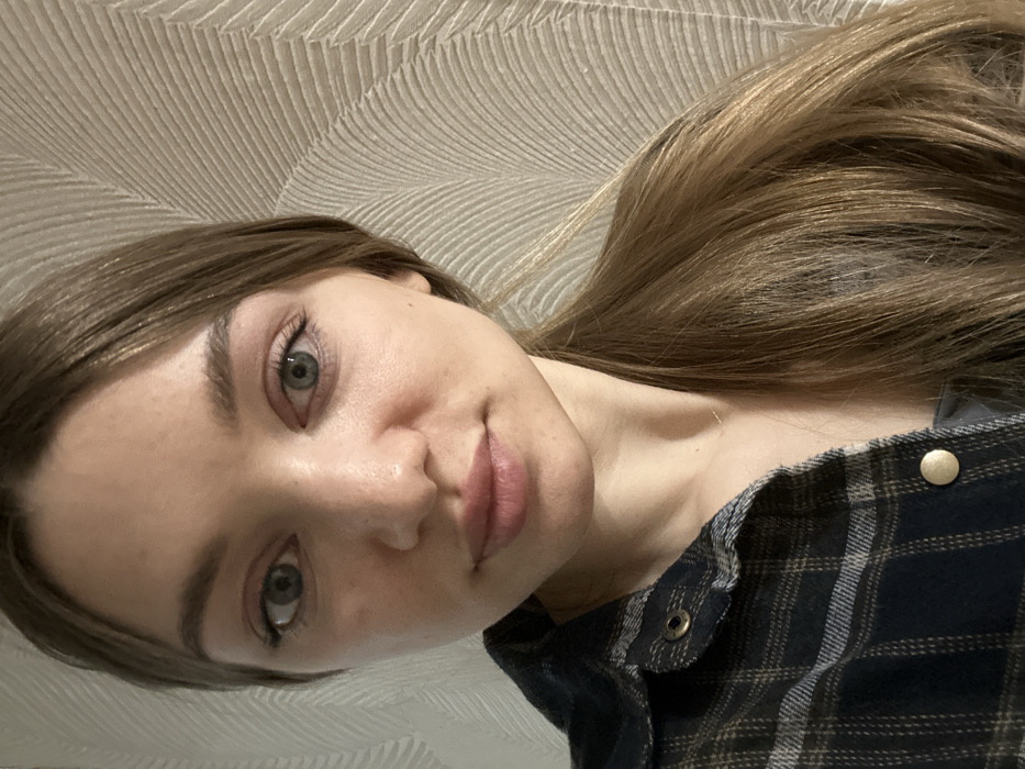 A close-up photograph depicts a female subject indoors, located in Япрыково, Russia. The subject faces the camera directly with a neutral expression. She has fair skin, light-colored eyes, and light brown hair pulled back or draped over her right shoulder. She is wearing a dark blue and beige plaid shirt with brass-colored snap buttons, unbuttoned to reveal a grey undershirt. A thin, gold-colored necklace is visible on her neck. The background consists of a light-colored wall with a textured, wave-like pattern.