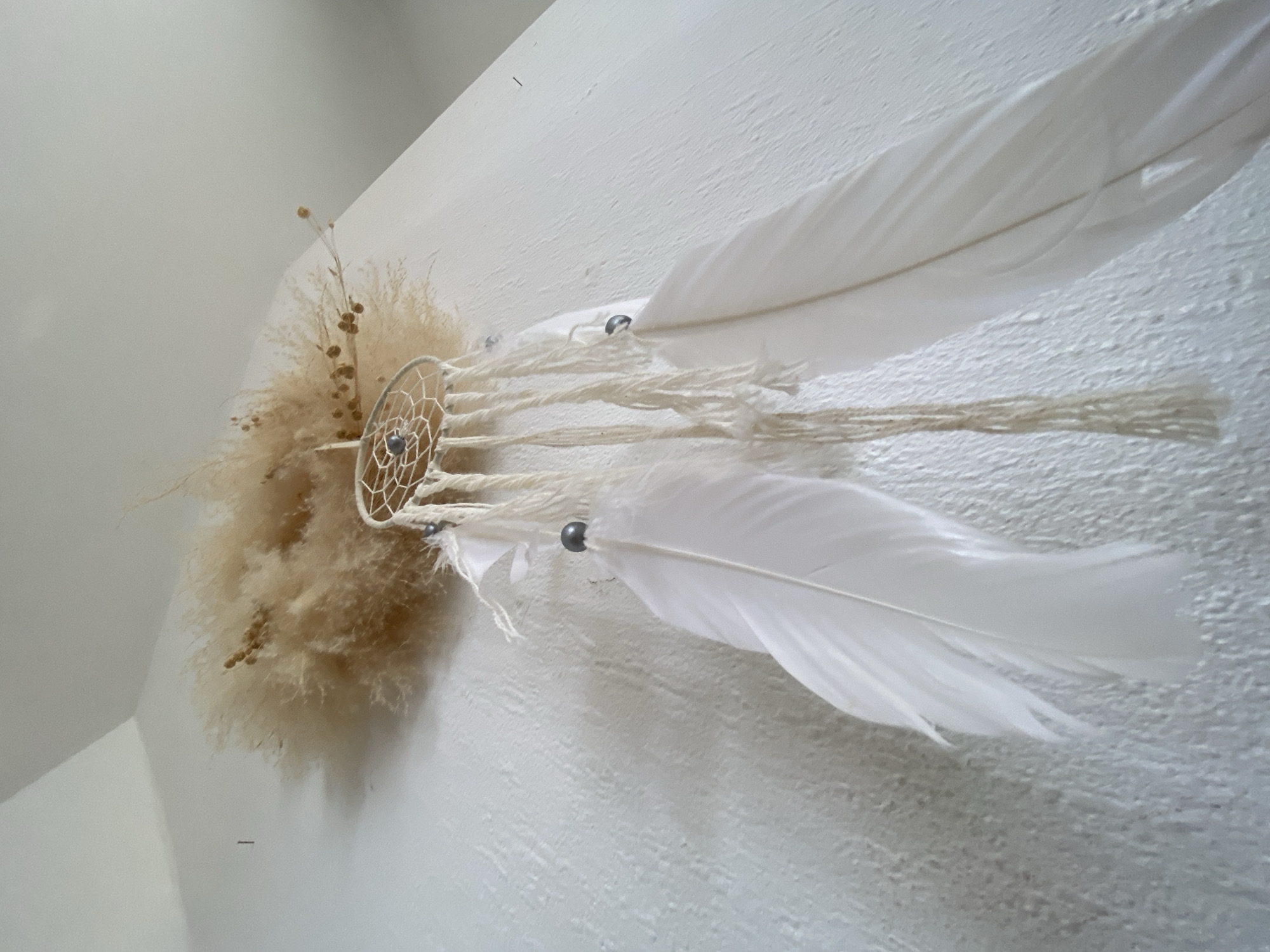A dreamcatcher is displayed against a white wall in a room, possibly in Bad Sachsa, Germany. The dreamcatcher is circular and woven with light-colored string, featuring a blue bead in the center. Dangling from the dreamcatcher are strands of cream-colored yarn with two large white feathers and several small gray beads. The background consists of a textured white wall and a sliver of a ceiling, suggesting an indoor setting.