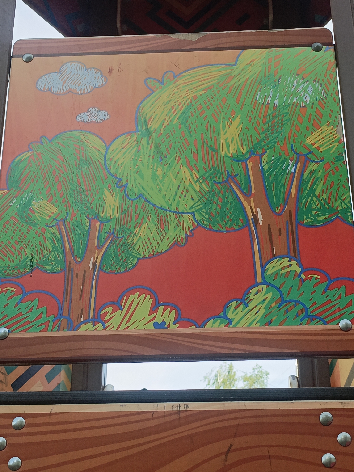 The image shows a section of playground equipment in Ufa, Russia. A painted panel depicting trees and clouds is framed by wooden supports. The panel's background is a gradient of orange to red, with green and yellow scribbled foliage and brown tree trunks outlined in blue. Two small white clouds are in the upper left corner. The playground structure is made of wood panels with visible grain and attached with silver bolts. Through a gap in the structure, a tree and sky are visible in the background.