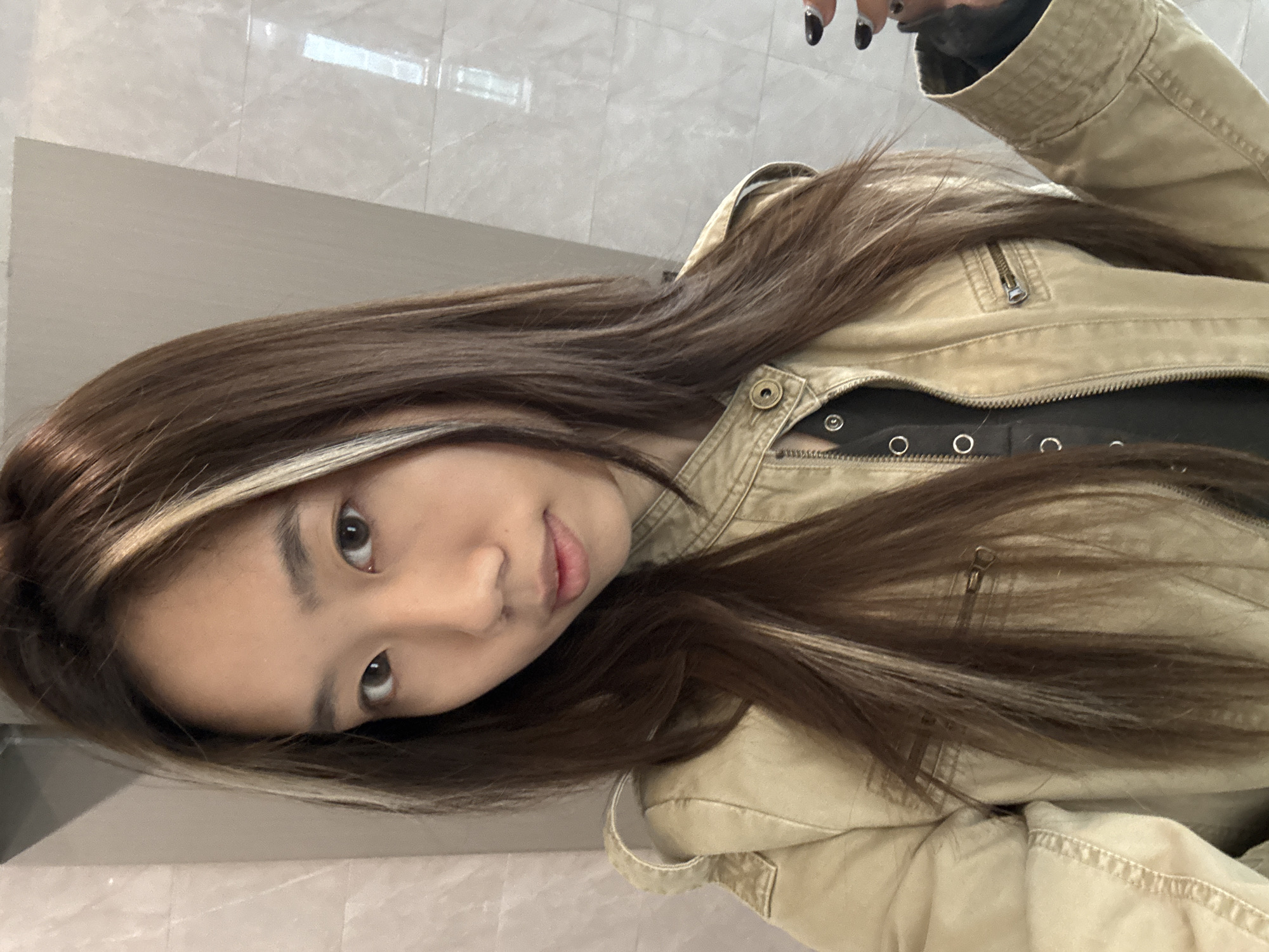 A young person with brown hair featuring a blonde highlight is wearing a tan-colored jacket over a black top with snap details. The background consists of light grey, possibly marble, wall panels or tiles. The image is rotated counter-clockwise. The location, Nan'an District, China, is not discernible from the image content.