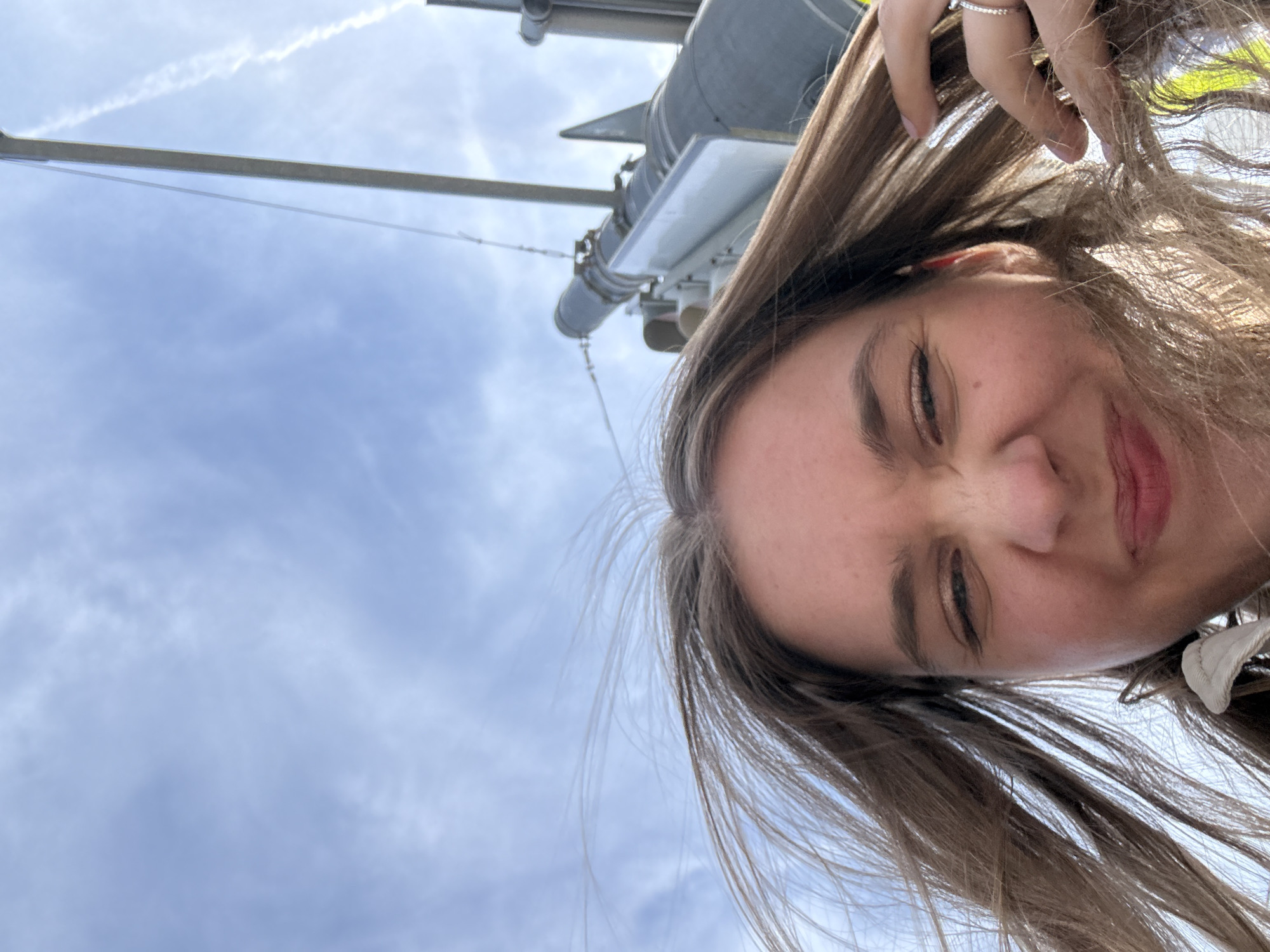 A tilted selfie shows a woman outdoors, possibly in Perm, Russia, given the query context. The sky is visible in the background, along with a pole and mechanical equipment. The woman is angled so her face is partially obscured by her hair. She is slightly squinting. She is wearing rings on her fingers. Her hair is brown.