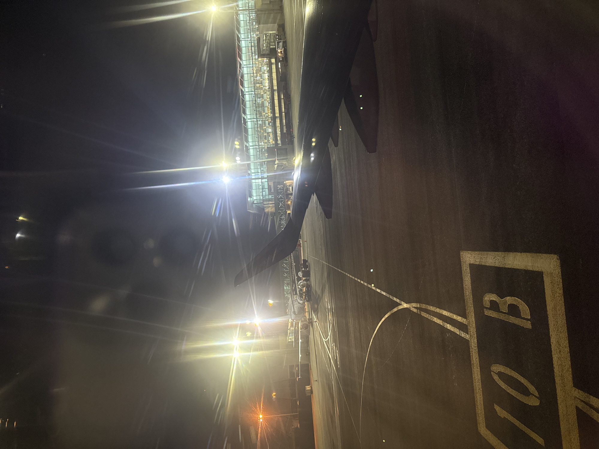This media file captures a nighttime scene at an airport, viewed from inside an airplane, likely through a window. The image is taken at an angle, showing the aircraft's wing prominently in the foreground, partially obscuring the view.
In the midground, a modern airport terminal building is visible, characterized by its glass and steel architecture, brightly illuminated from within and by exterior lights. These lights create strong lens flares and starburst effects across the dark sky, particularly on the left side of the frame. Below the terminal, various ground support equipment, including what appears to be jet bridges or mobile stairs, are positioned near the aircraft. Several small figures, likely ground crew, can be discerned near the wing and equipment.
The tarmac in the foreground and midground is dark, contrasting with the bright lights. It appears somewhat wet or reflective in areas. Clear white and yellow markings are painted on the asphalt. A large, prominent marking "10 B" is clearly visible in the lower right corner, likely indicating a gate or stand number. Other faint lines and curves, typical of airport ground markings, are also present.
The overall scene depicts an airplane parked at a gate or preparing for departure/arrival at an airport in Yerevan, Armenia, during the dark of night. The strong artificial lighting against the darkness emphasizes the industrial and functional environment of the airport at night.