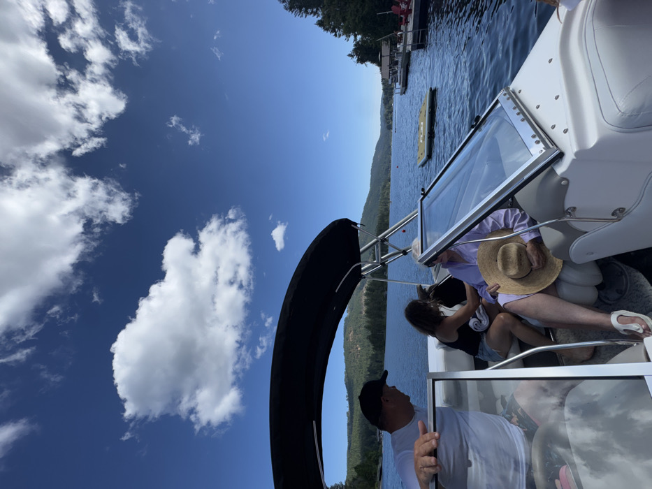The image depicts a daytime scene on Shawnigan Lake in Area B, Canada, from the vantage point of a white and grey pleasure boat. Three individuals are present on the boat. In the left foreground, a male wearing a white short-sleeved shirt and a dark baseball cap leans with his right arm on a boat frame, looking towards the center. In the mid-ground, a female with dark hair, dressed in a dark tank top and light shorts, is seated and appears to be attending to another seated female beside her. This second female, older in appearance, is wearing a light purple long-sleeved garment and white flip-flops, holding a wide-brimmed straw hat on her lap. A black bimini top extends over the boat's seating area, and a clear, framed windshield is visible on the right side of the boat, angled upwards.
The boat is positioned on calm, blue lake water. To the right, a yellow inflatable mat floats near a wooden dock adorned with red chairs on the tree-lined shoreline. The background features rolling forested hills encircling the lake under a bright blue sky dotted with scattered white cumulus clouds. Several other boats are visible in the distance on the water, along with a red buoy.