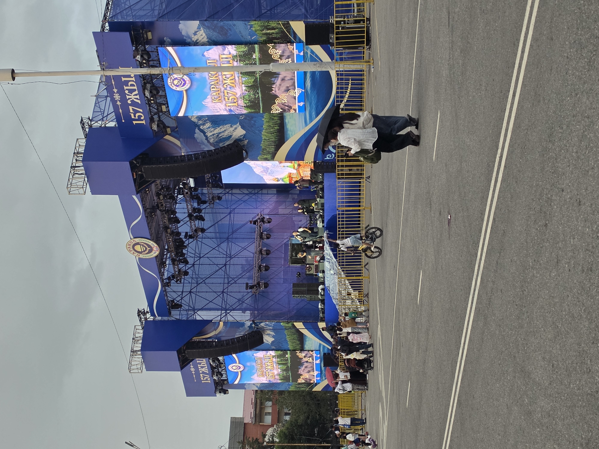 The image depicts an outdoor event in Karakol, Kyrgyzstan, likely a celebration or festival. A large, elaborate blue stage with multiple screens dominates the background. The screens display images of mountains and text, including "157 ЖЫЛ" and "КАРАКОЛ 157 ЖЫЛ". Several speakers are mounted on the stage structure.
In the foreground, a group of people are gathered, some standing and some sitting behind a yellow barrier. Two individuals in the middle ground are under a black umbrella, appearing to interact closely. Further to the right, a person is performing a stunt on a bicycle, with one wheel in the air. The ground is asphalt with white lane markings.
The weather appears overcast, suggesting it might be a cloudy day. The presence of a large stage and a gathering of people indicates a public event. The text on the screens suggests that the event might be a celebration of 157 years of Karakol. The overall mood seems festive, with the bicycle stunt adding an element of excitement and performance. A few buildings are visible in the distance on the left side of the image.