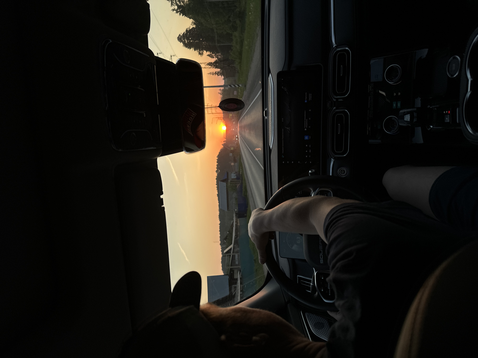 The image depicts the interior of a car driving on a road towards the sunset. The sun is visible in the distance, shining brightly down the road. The dashboard of the car is visible, as well as the steering wheel with a hand holding it. The view out the windshield shows trees and buildings lining the road, typical of a rural area. The road is likely located within the city of Chaikovsky, Russia, given the location information. The photo is rotated, requiring a 90-degree rotation to view correctly.