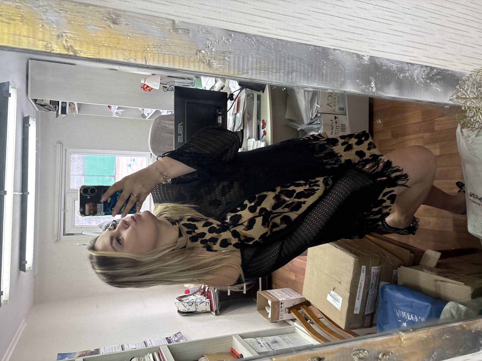 A blonde woman is taking a selfie in a mirror in what appears to be a cluttered office or storage space. She is wearing a black mesh dress with fringe details and a leopard print scarf around her neck. She is puckering her lips and holding her phone in her right hand. The background shows shelves filled with boxes, papers, and office supplies. There is a window with green blinds visible behind her. The floor is wooden. The overall impression is one of a candid, informal moment captured in a utilitarian environment. The location is identified as Noviy, Russia.