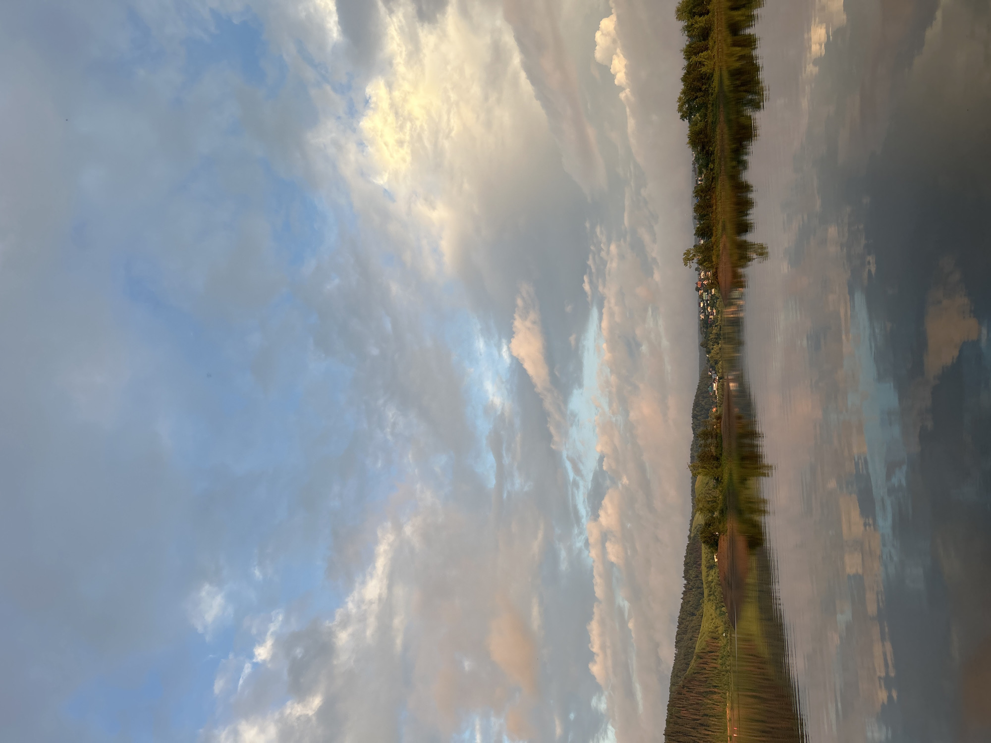 The image depicts a wide, calm river reflecting a cloudy sky and a tree-lined shore. The sky is a mix of blue and grey clouds, with patches of golden light, suggesting either sunrise or sunset. The shore features rolling hills covered in dense green forest, with a small cluster of buildings visible in the distance on the right side of the frame. The water is still, creating a mirror-like reflection of the sky and the landscape. There are no people or animals visible in the image. The scene is serene and natural, with no indication of urban activity beyond the distant buildings. The location is identified as the city of Noviy, Russia.