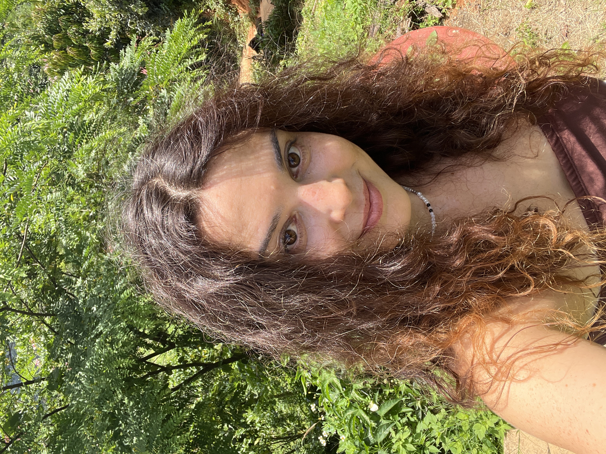 A woman with curly brown hair is taking a selfie outdoors. She is wearing a brown top and a beaded necklace. She is smiling at the camera. The background is lush and green with trees and foliage. It is daytime and sunny. The image does not provide enough information to determine the exact location within Sơn Điền, Vietnam.