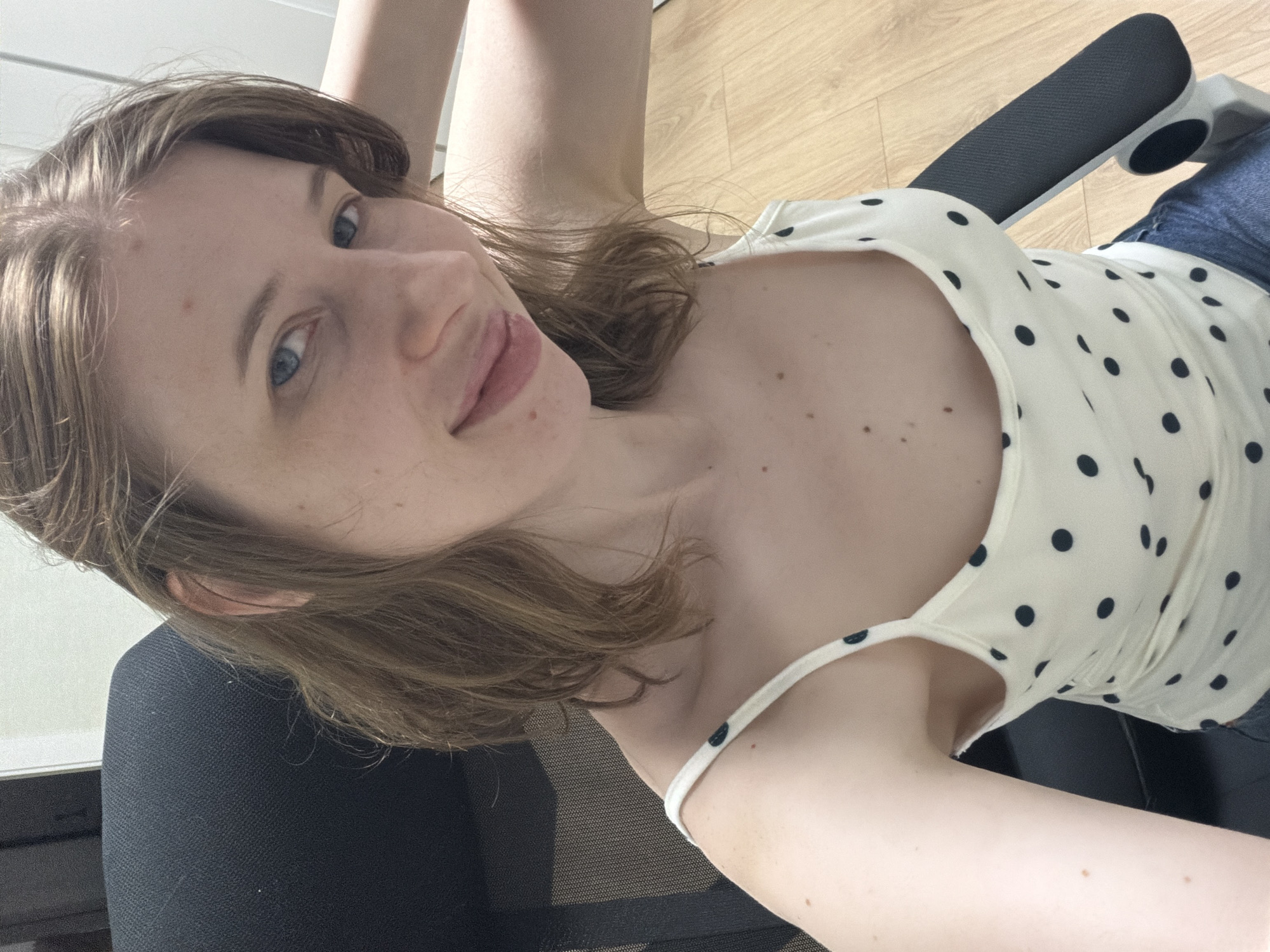 A young woman with light brown hair and blue eyes is seated in a black office chair. She is wearing a white tank top with black polka dots and blue jeans. Her right arm is bent behind her head. The background appears to be an indoor setting with light-colored walls and a wooden floor. There is no indication of the location being in Dongducheon-si, South Korea.