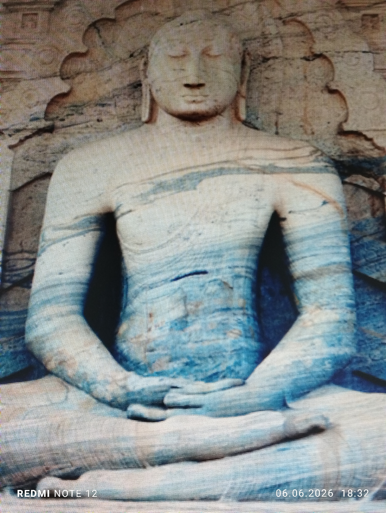 The image presents a photograph of a digital screen displaying a carved stone statue of a seated Buddha. The statue, appearing to be crafted from light-colored stone, exhibits prominent horizontal and wavy blue-grey patterns or striations across its surface, particularly on the torso, arms, and legs. The figure is depicted in a meditative posture, seated cross-legged, with both hands clasped in the lap in a dhyana mudra. Essential facial features include closed eyes, a serene expression, elongated earlobes, and a cranial protuberance (ushnisha). The background behind the statue's head and shoulders is also carved, featuring decorative arch-like or flame-like motifs. The overall image quality is pixelated, consistent with a photograph taken of a digital display. The location of the scene is in Burgas, Bulgaria. No individuals or dynamic actions are present. Timestamp "06.06.2026 18:32" and device information "REDMI NOTE 12" are visible in the lower right and left corners, respectively.