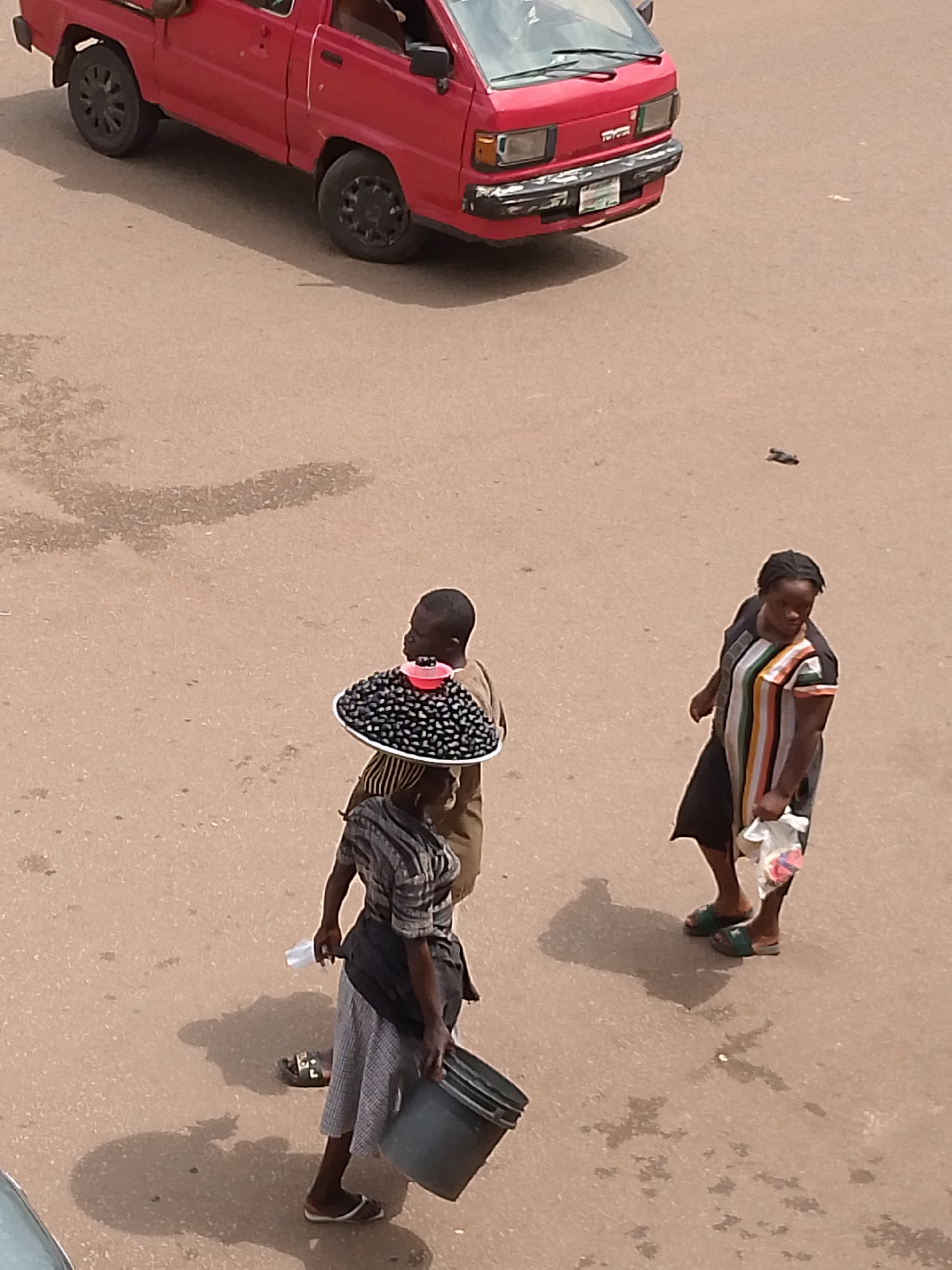 In Bukuru, Nigeria, a red Toyota pickup truck is parked on a paved surface. Three individuals are present. One person, a woman wearing a striped dress, stands with a plastic bag in her right hand and a white object in her left. Another person, a woman in a patterned skirt and striped top, carries a black bucket in her right hand and a white cup in her left. A third person, facing away from the camera, is carrying a circular tray on their head filled with dark, small, round items, topped with a pink bowl. The ground is a dusty brown asphalt with some scattered debris. Shadows are cast on the ground, indicating a sunny day.