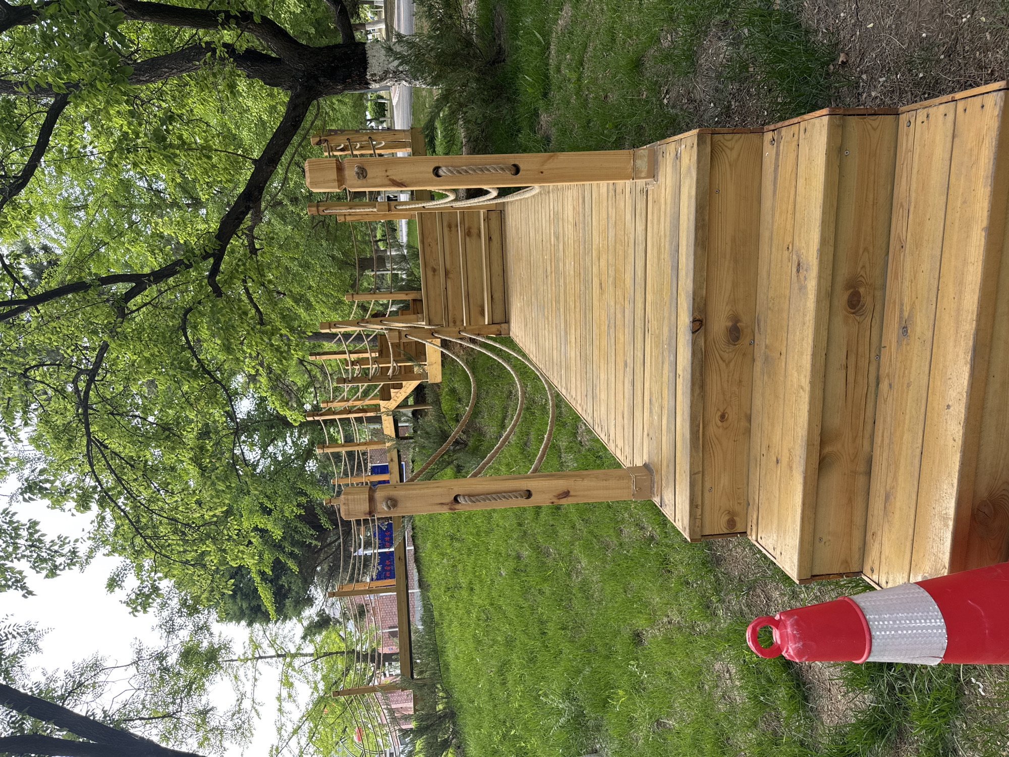 The scene depicts a wooden walkway structure in what appears to be a park or recreational area. The structure is constructed of light-colored wood, featuring a deck-like platform with railings made of rope. The ground surrounding the structure is covered in green grass. To the side of the structure, a red and white traffic cone lies on the grass. The background features a blurred view of trees, suggesting a park environment. According to the image metadata, the location is the city of Zhongtun, China. The image provides no details about people, specific actions, or interactions.